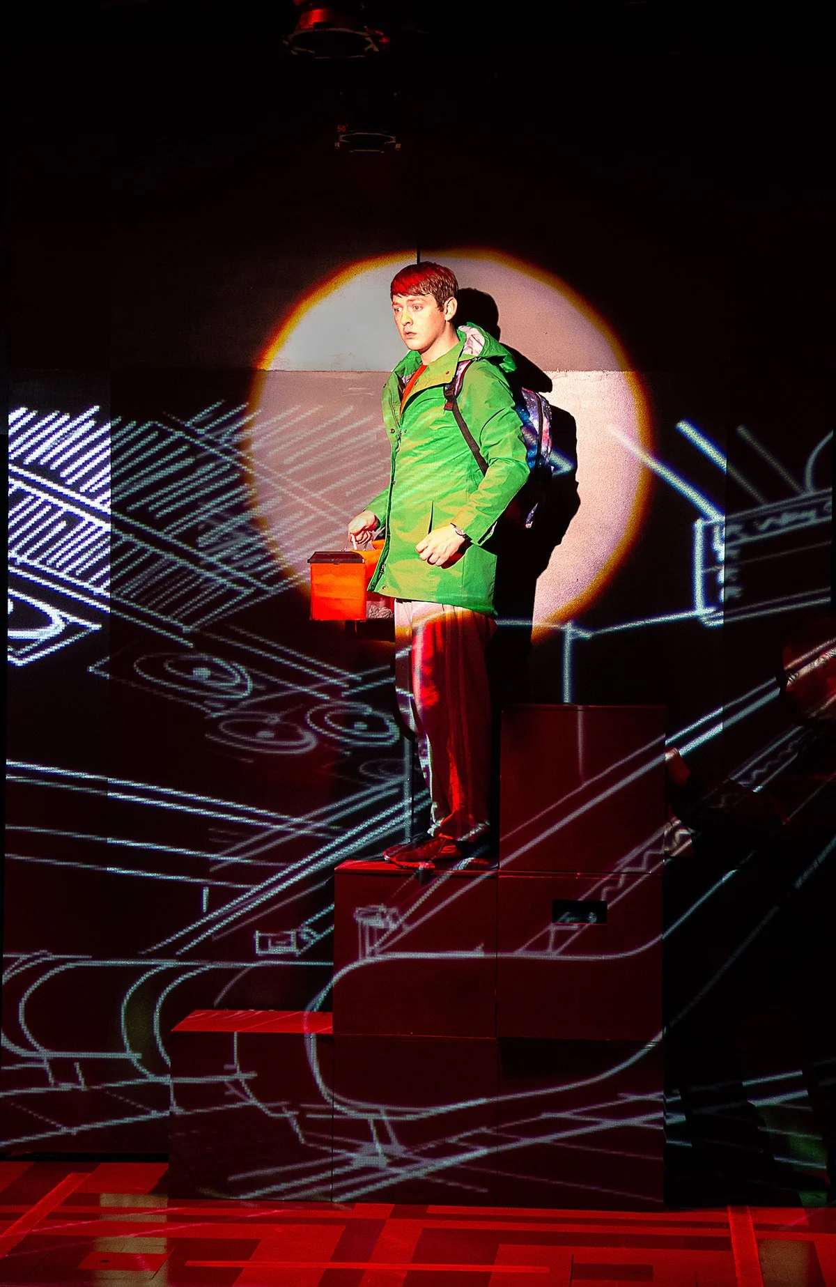 A young man in a green jacket with a backpack, holding a briefcase, standing on a red podium against a background of projected architectural blueprints and a circular light.