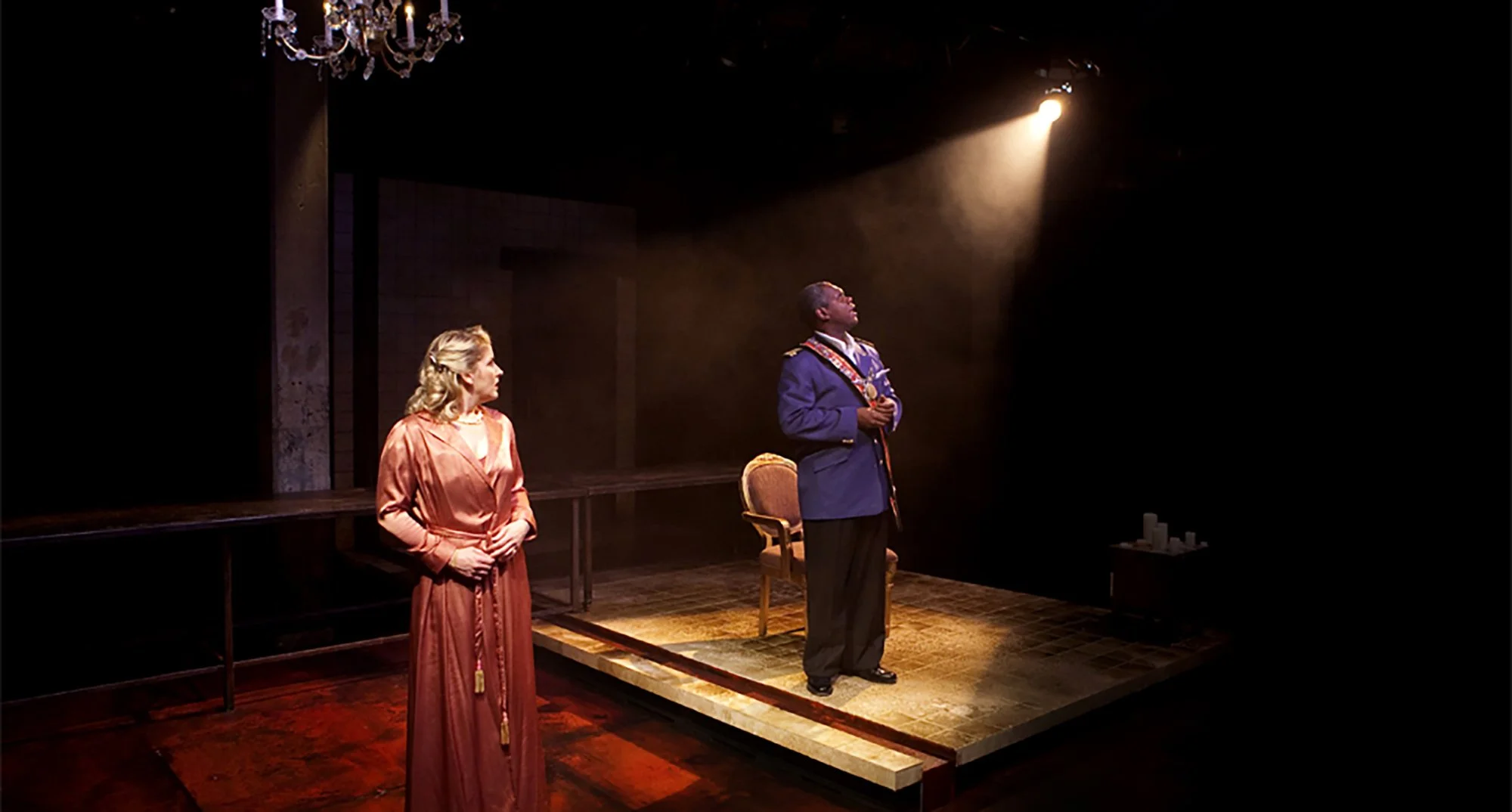 Two actors on stage, a woman in a pink dress looking at a man in a military uniform who is standing with hands clasped, illuminated by stage spotlight.