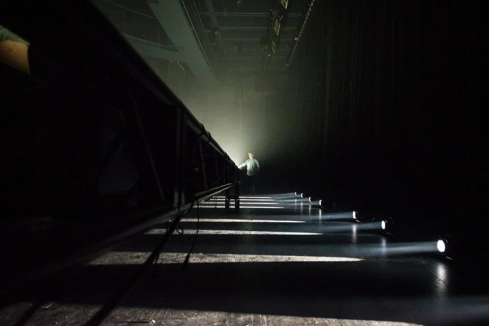 A person standing on a dark stage illuminated by a bright white spotlight, with several small ground lights lining the edge of the stage.