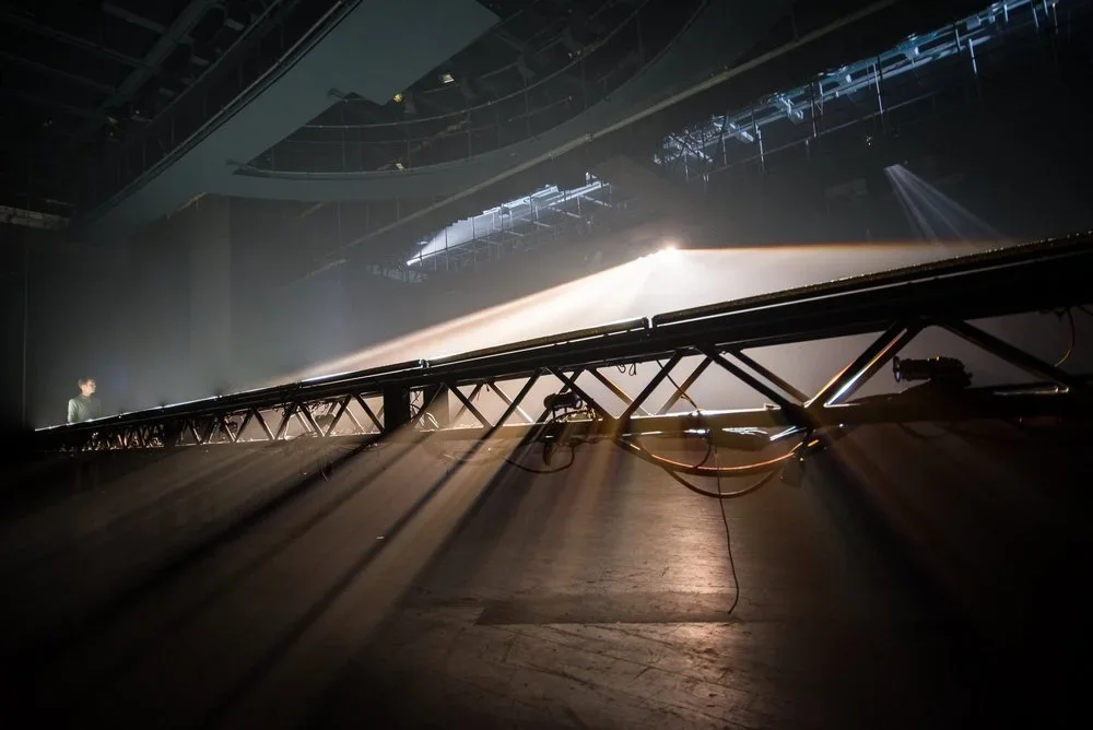 Stage with lighting equipment and a crew member, bright spotlight shining on the stage, possibly during concert or performance setup.