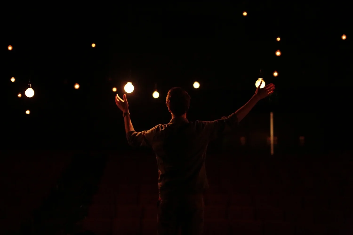 A person standing with arms outstretched amidst hanging light bulbs in a dark space.