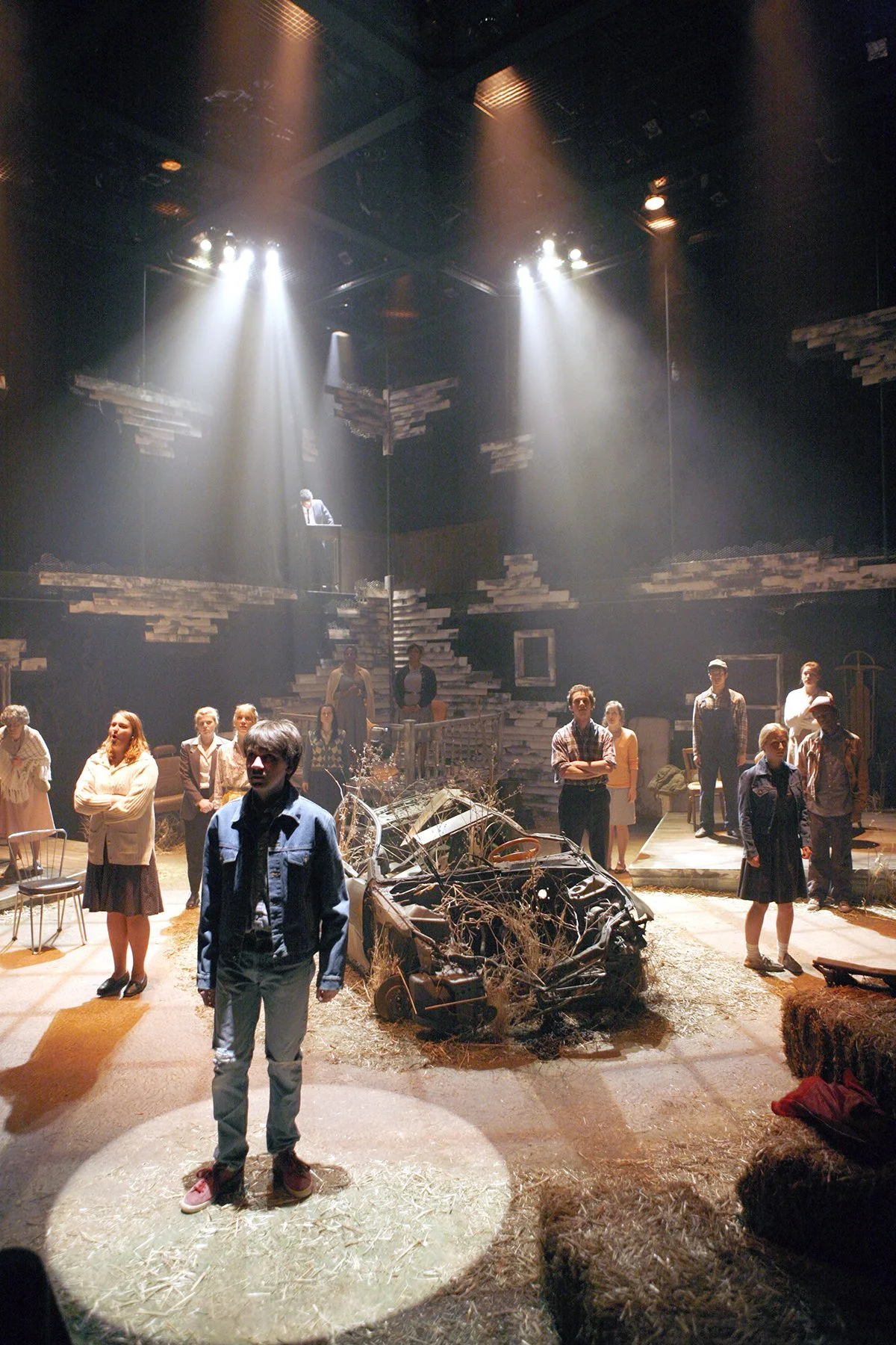 The image shows a theatrical stage scene with a group of actors, some standing and some sitting, around a damaged car with branches and debris on it. The stage set includes a wall made of uneven stacked wooden planks and a small elevated platform at 
