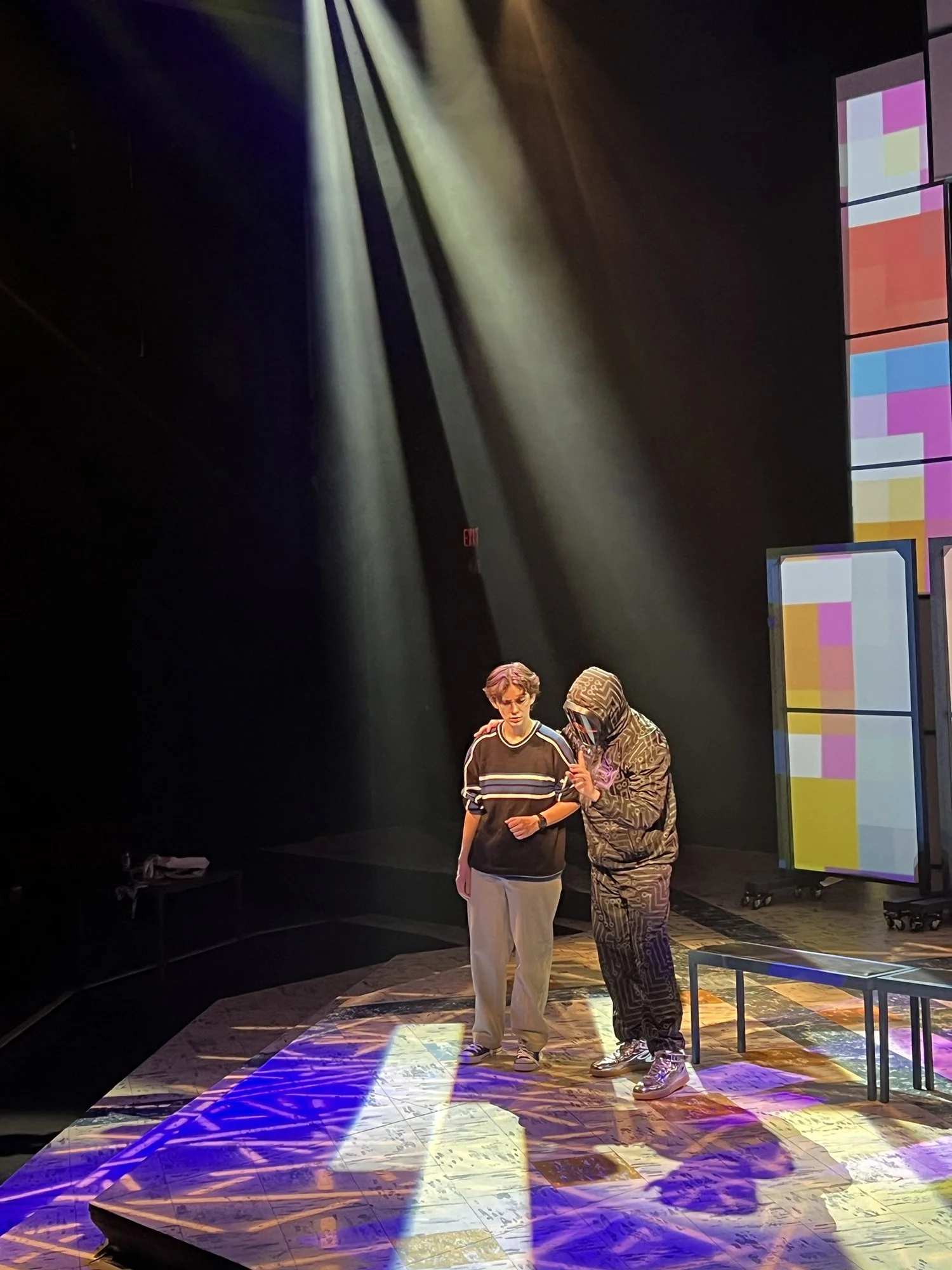 Two people, a man and a woman, on a stage illuminated by colorful lights. The man is wearing a striped t-shirt and beige pants, while the woman is dressed in a patterned hoodie and matching pants. A multimedia screen displaying colorful blocks is vis