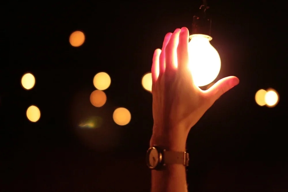 A hand reaching up to touch a glowing light bulb against a dark background with out-of-focus lights.