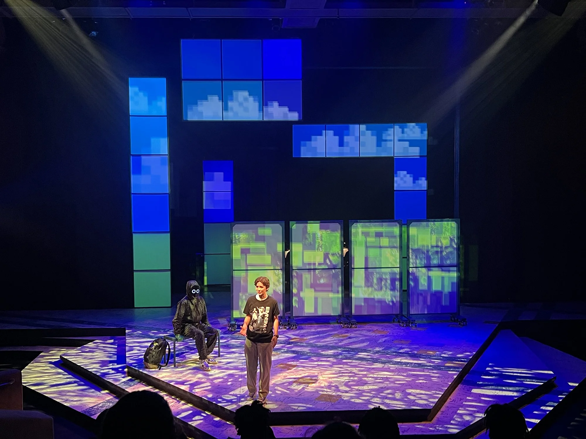 The image shows a stage with two people sitting and one standing in front of a colorful, pixelated digital art display. The background features numerous blue and purple illuminated pixel art panels, some depicting pixelated clouds. The stage is lit w