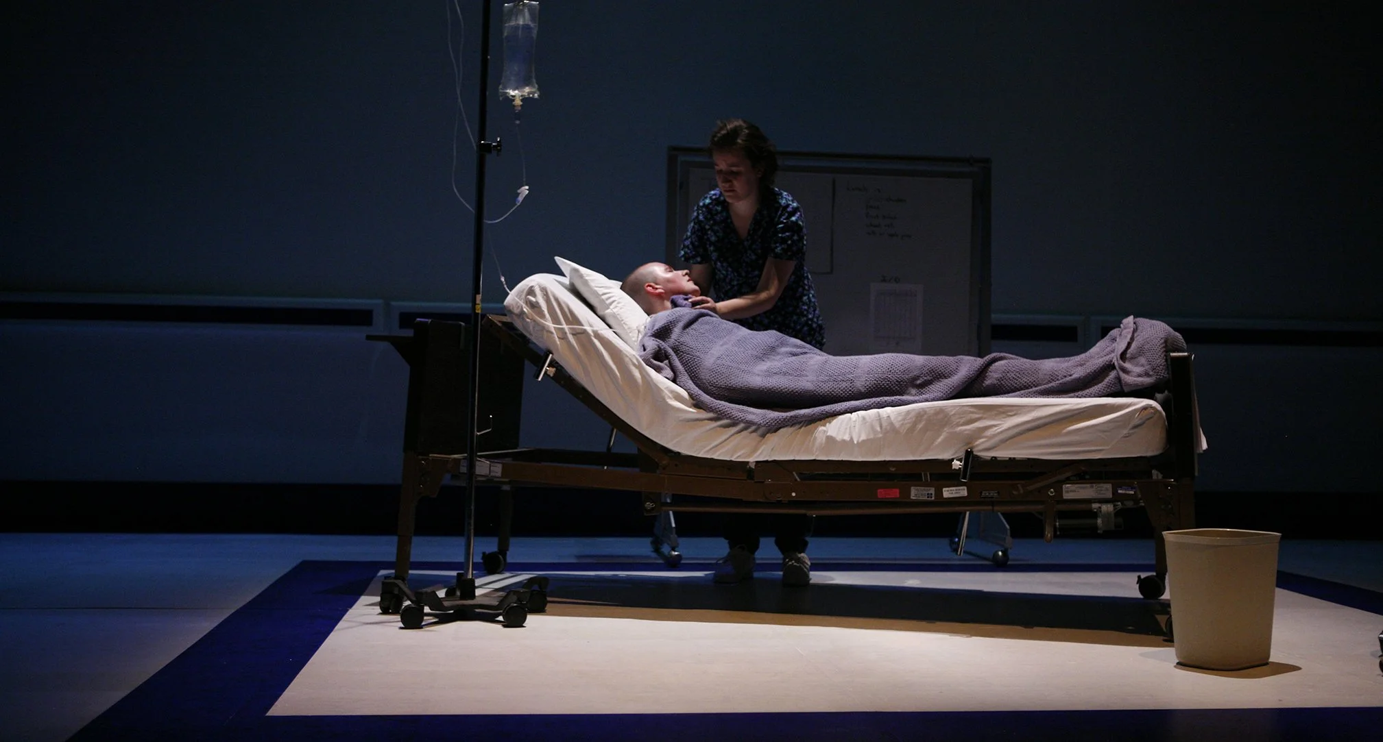 A person lying in a hospital bed with an IV attached, covered with a gray blanket, and another person tending to them in a dimly lit room.