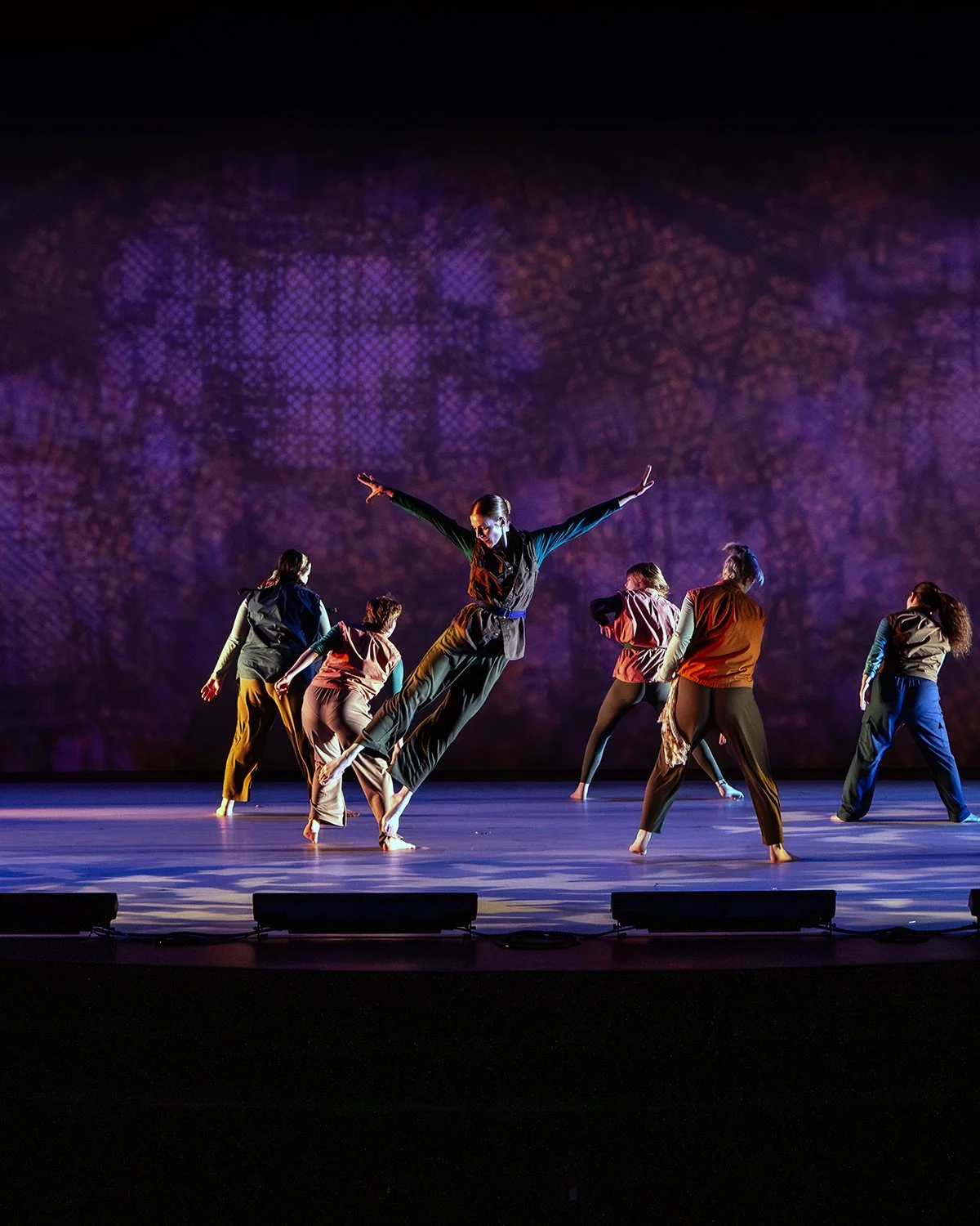 Dance performance with six female dancers on stage, colorful costumes, purple-lit background.