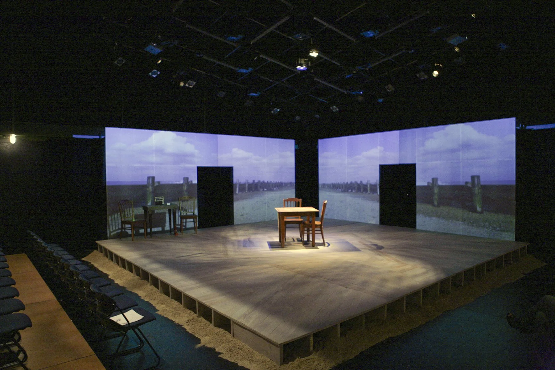 Theater stage with two screens displaying a seascape with clouds and wooden pier posts, and furniture including chairs and a table on the stage.