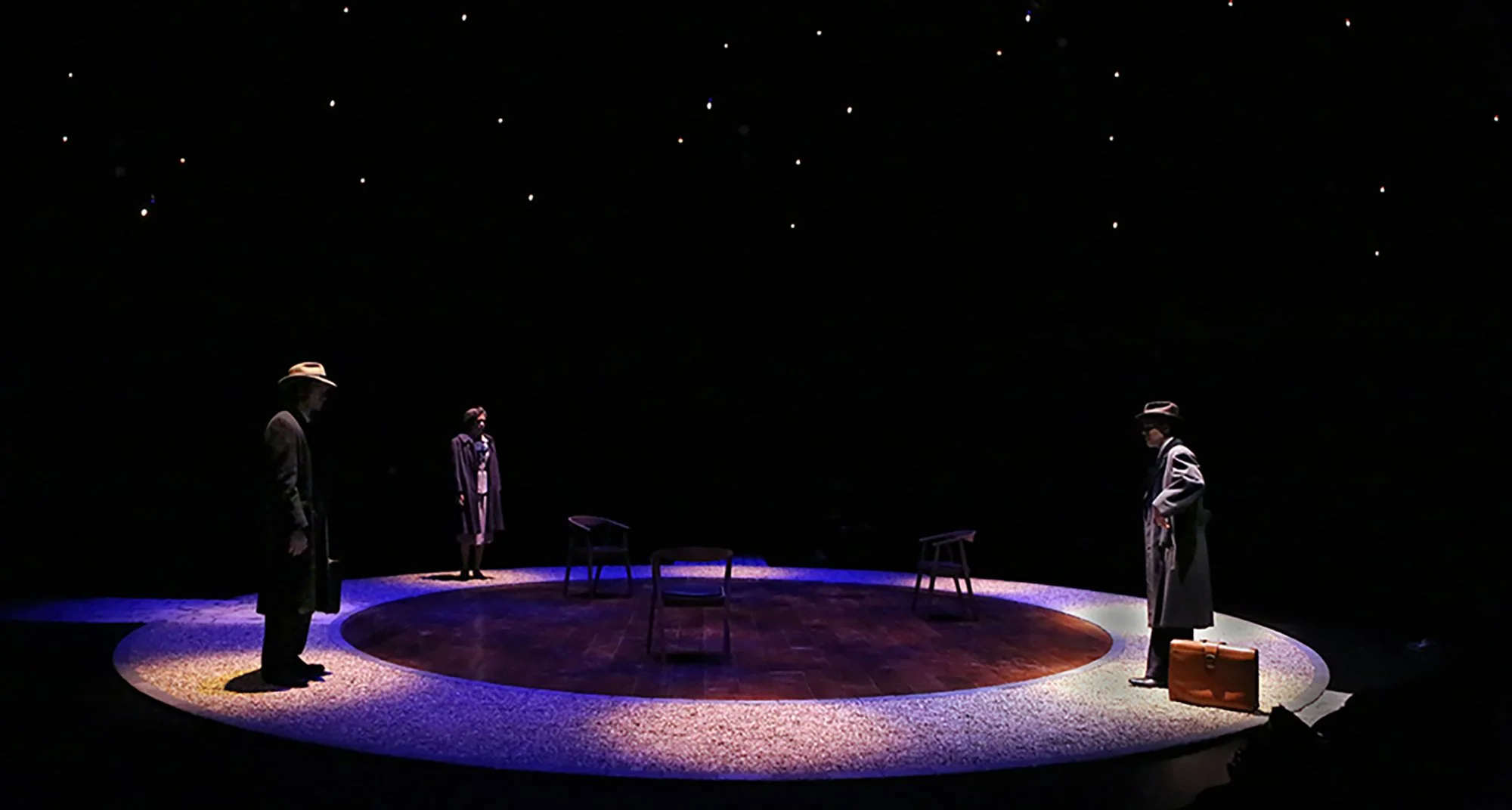 Three actors in costume standing on a circular stage with minimal furniture, under dim lighting with small star-like lights overhead.