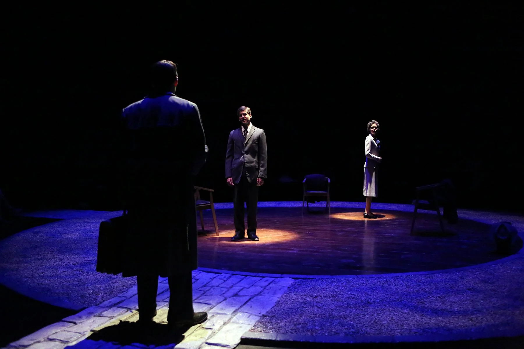 Three people standing on a circular stage in a dark setting; two are illuminated by spotlights, and one person appears in the foreground with their back to the camera. The background is mostly dark with a few chairs visible around the stage.