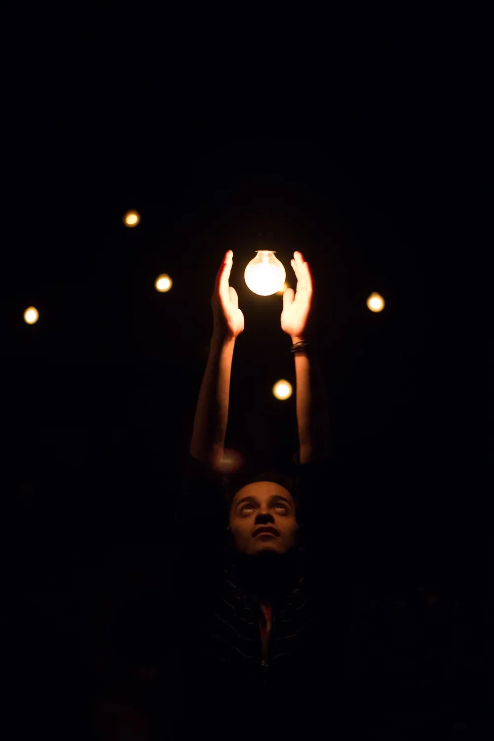 Person holding a lit light bulb above head in darkness, surrounded by small blurred lights.