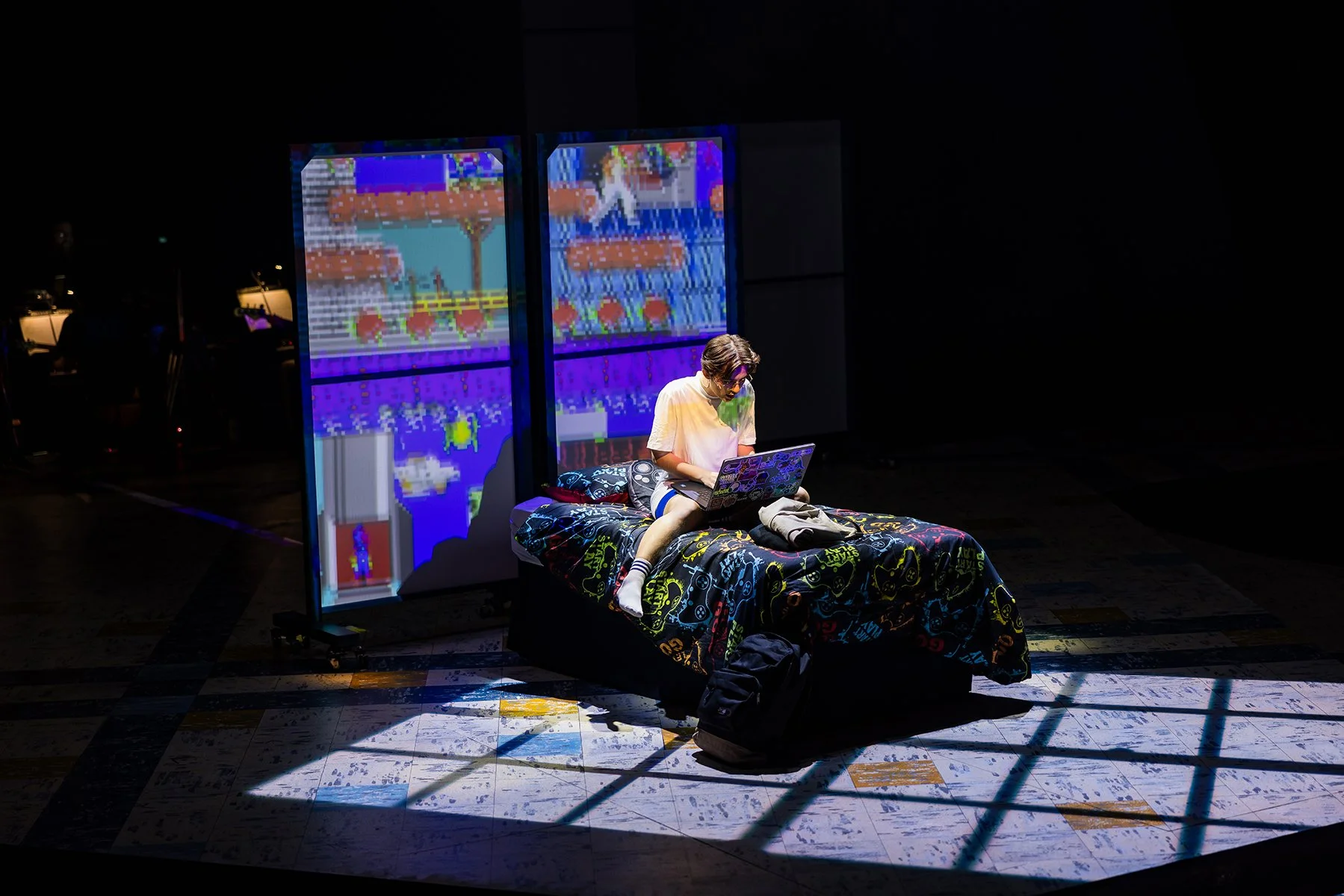 A person sitting on a bed with a colorful blanket, using a laptop, in front of two large pixelated screens displaying video game graphics, in a dark room.