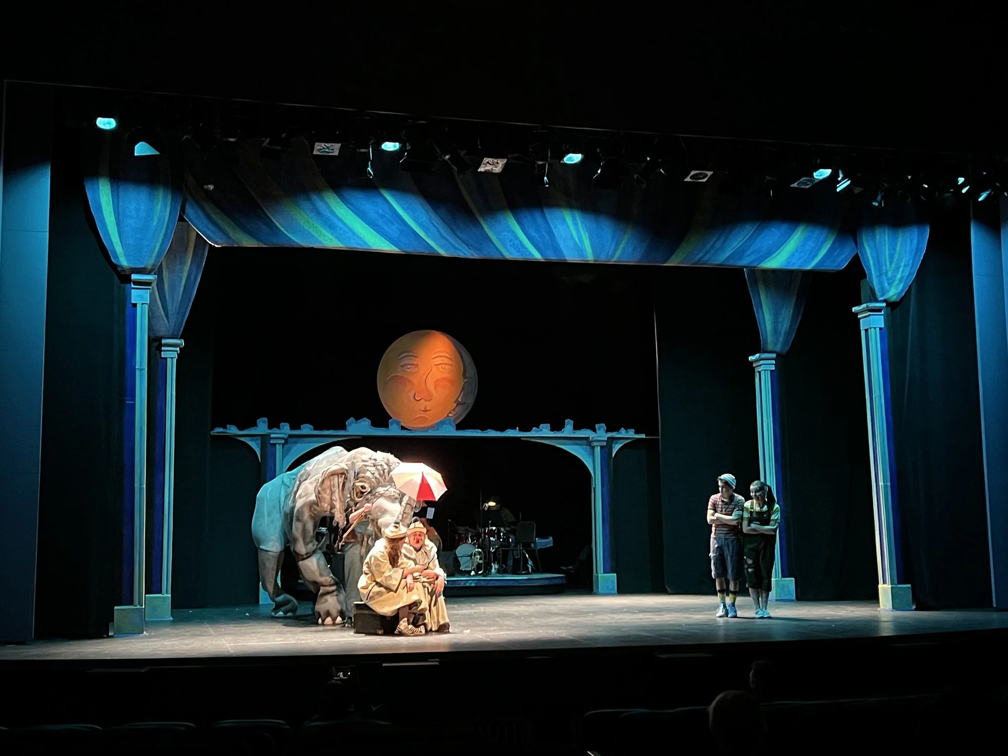 A stage setup with a theatrical performance featuring actors in costume, including an elephant and a person sitting on a throne, under a blue and green curtain, with stage lights overhead and a decorative face in the background.