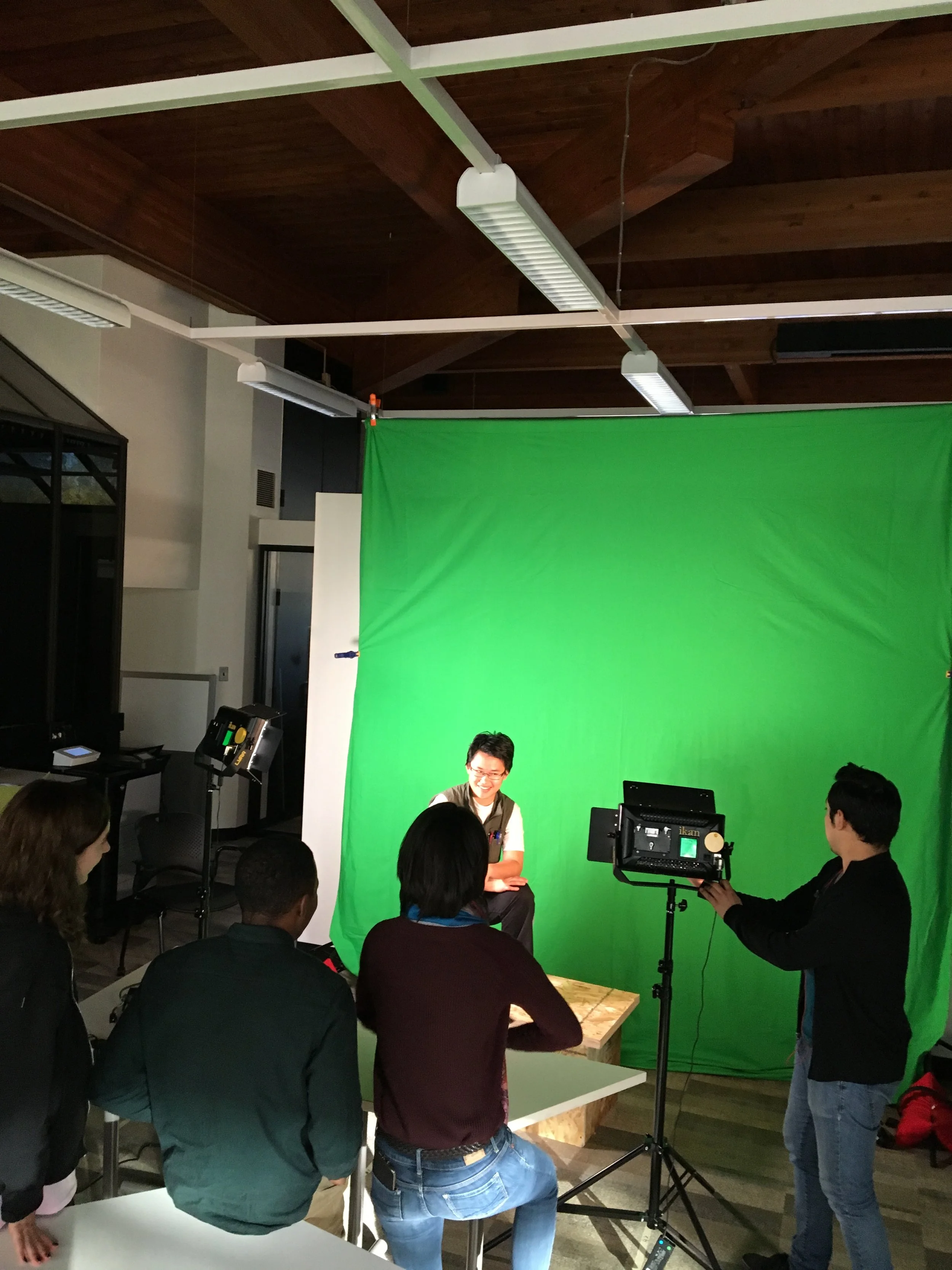 Group of people in a room with a green screen, some sitting and some standing, with equipment and a person operating a camera.