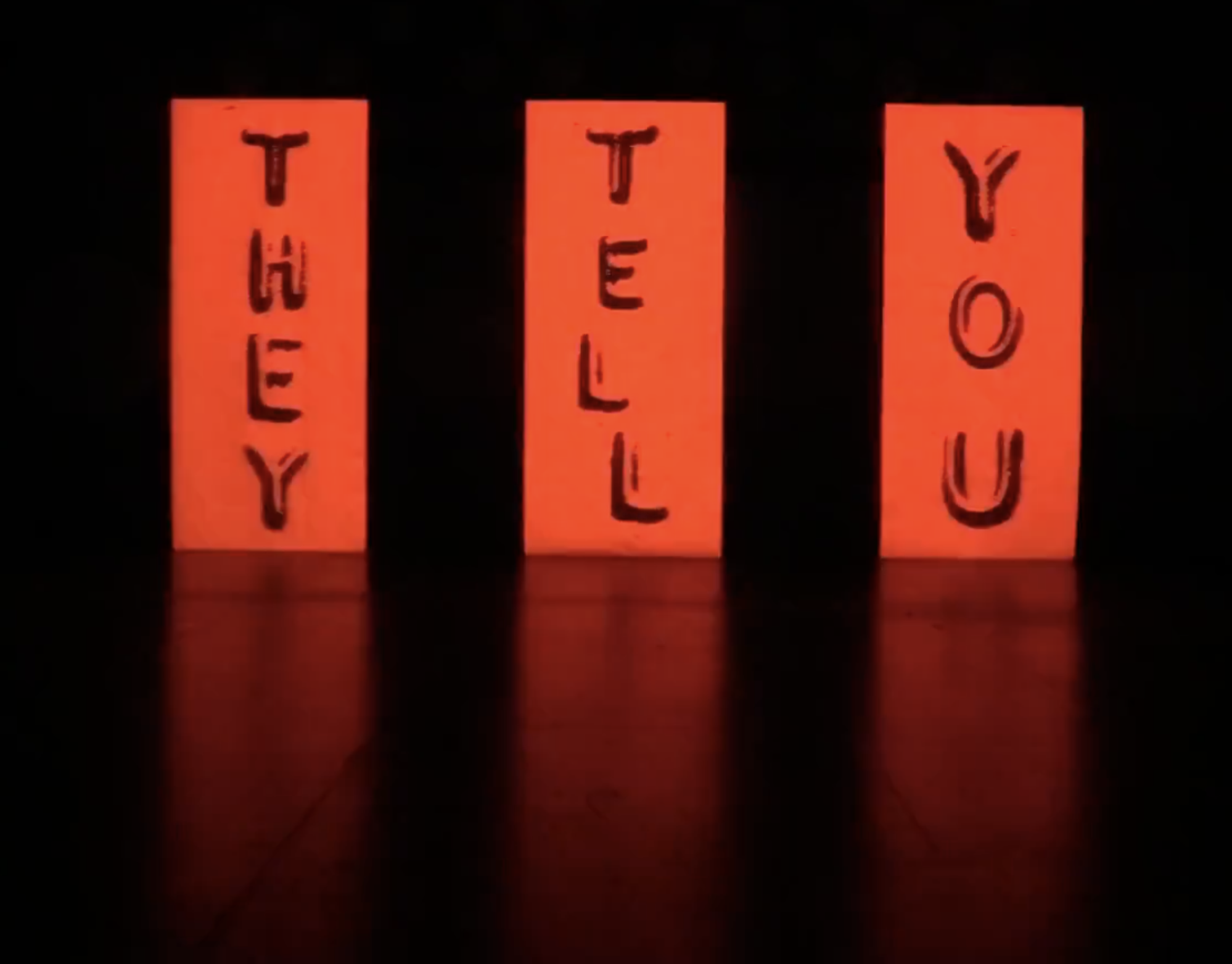 Three glowing red neon signs with the words 'HEY,' 'TELL,' and 'YOU' written vertically on black background.
