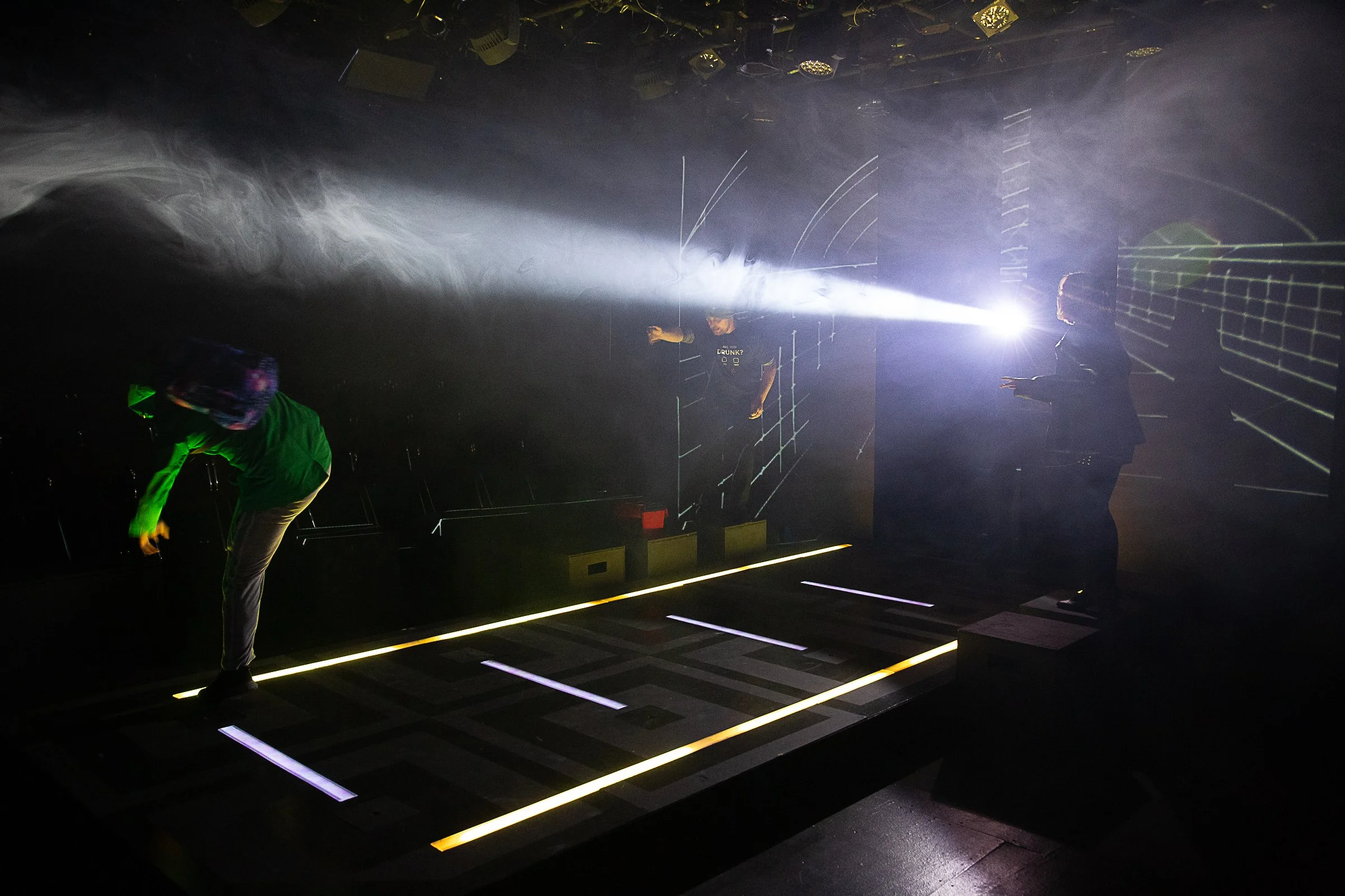 People on a dark stage with laser light beams and grid patterns projected onto the floor and background.