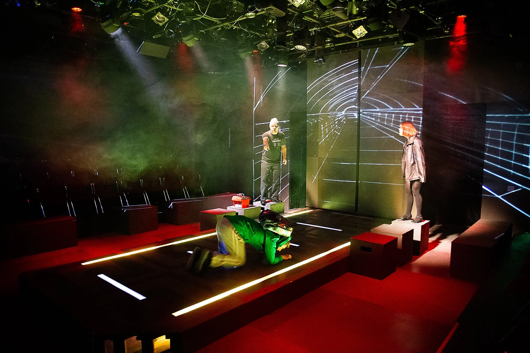 A theater stage with digital graphics projected on the background, three performers on stage, one lying on the floor, one standing on a platform pointing, and one standing on a raised block, with colorful lighting and set pieces.
