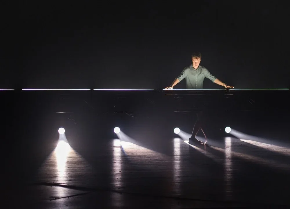 A person standing on a dark stage or dance floor, illuminated by four bright spotlights casting beams of light on the floor, with one foot slightly lifted, doing a dance or performance.