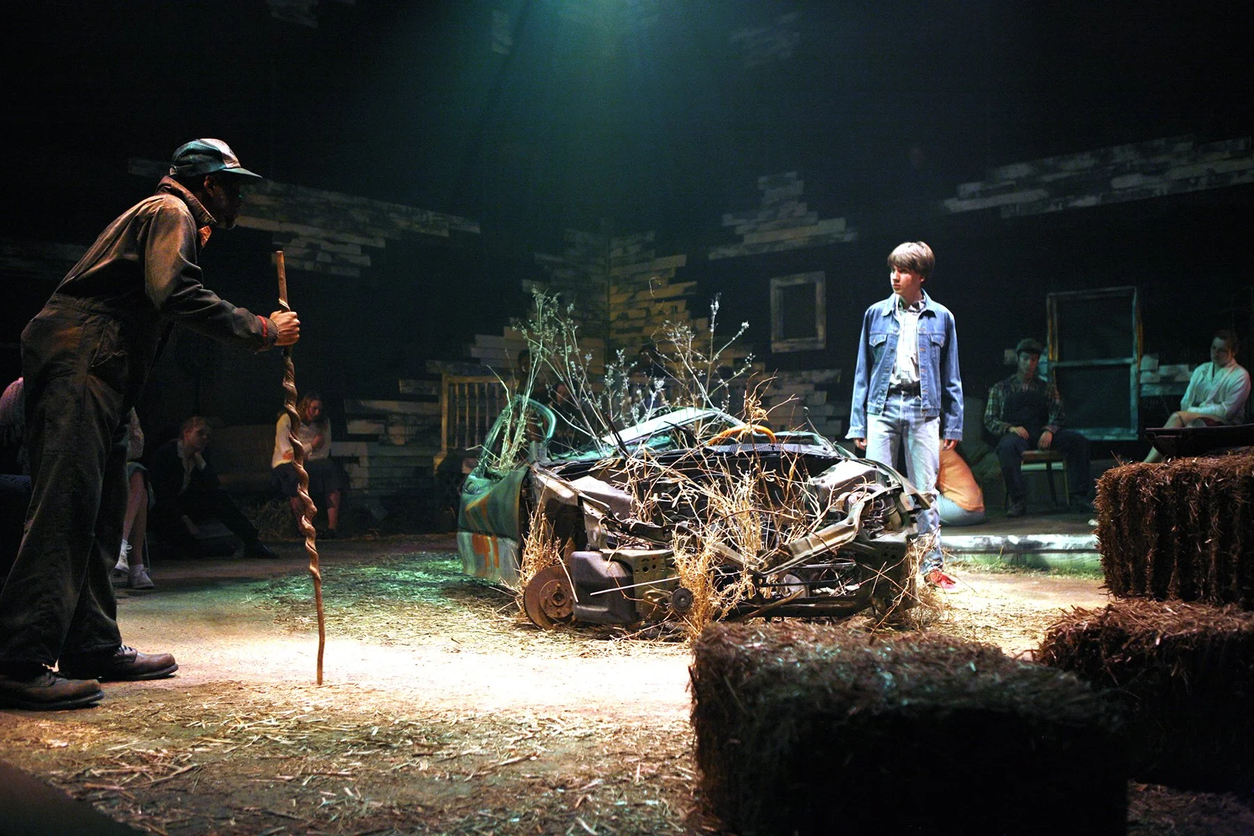 A theatrical performance depicting a scene with a damaged car, dry plants, and actors, including a young boy in a denim jacket, on a stage with wooden walls and seated audience members.