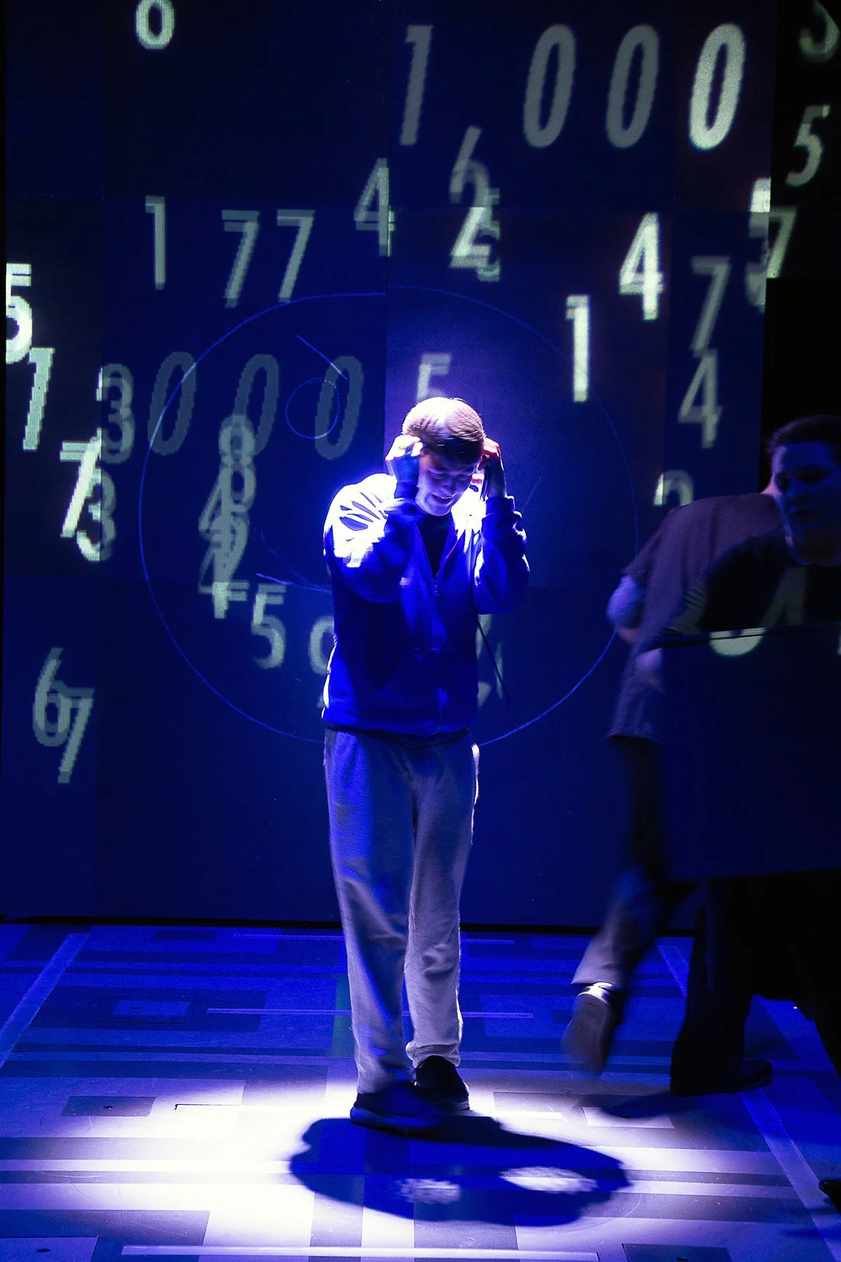 A young person standing in front of a dark background with large glowing numbers and circles projected behind them. They are wearing casual clothing and appear to be adjusting their headphones or accessories.