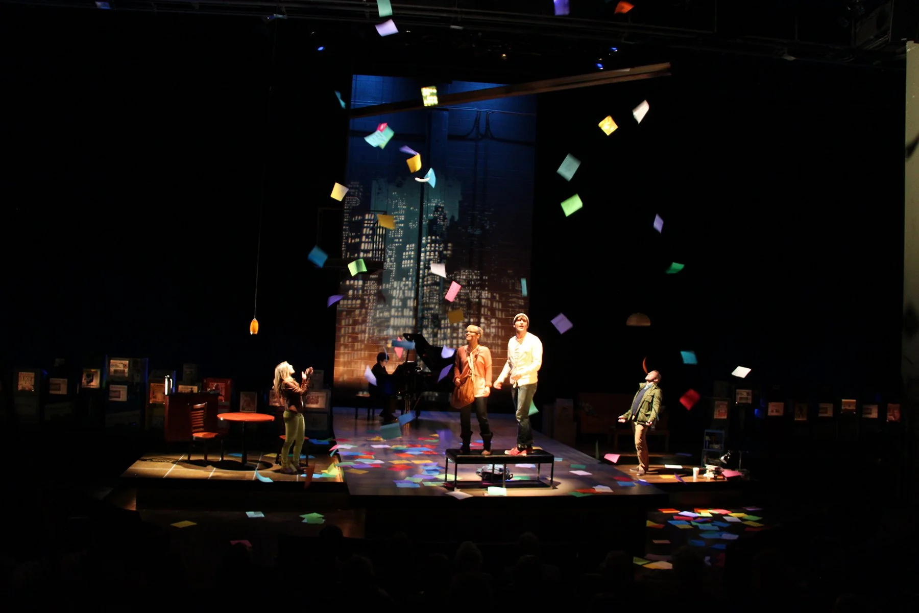 The image shows a theater stage with actors performing under dark lighting, with colorful papers falling from above. In the background, there is a cityscape projected on a screen. The stage is decorated with furniture and props, including chairs, tab