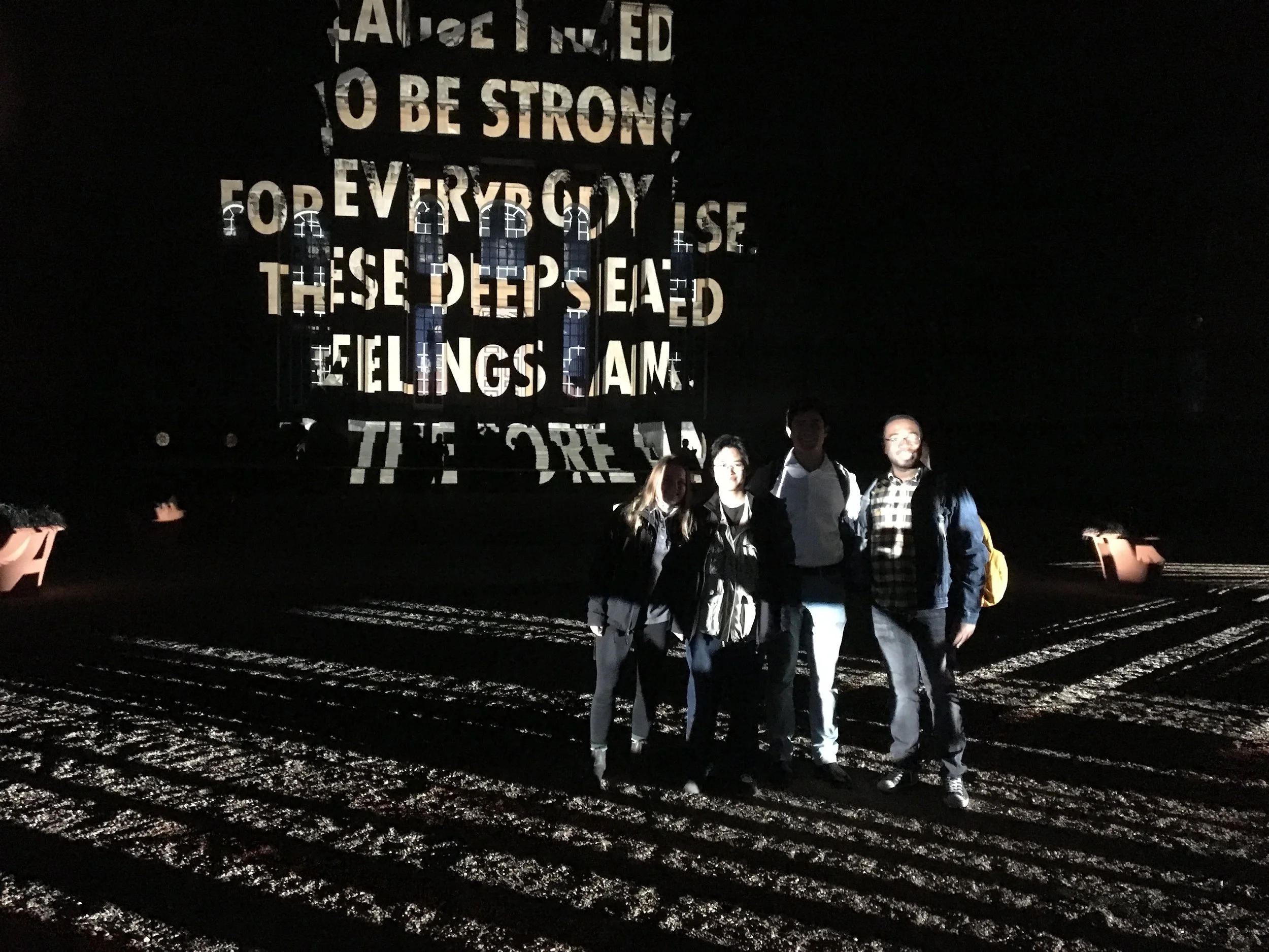 A group of five people standing outdoors at night with a dark background and an illuminated sign displaying a partial quote. The ground is wet and reflects light.