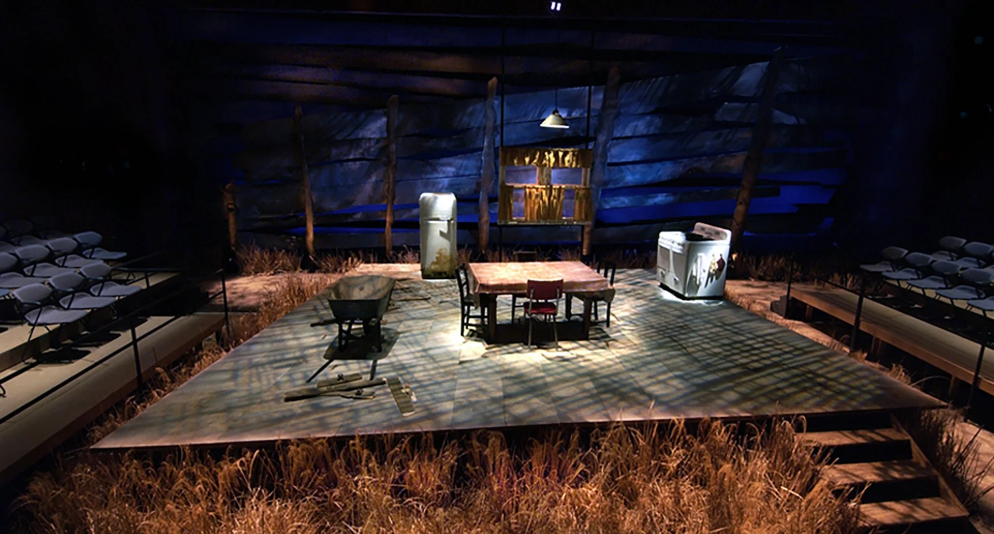 A theatrical stage designed like a rustic outdoor scene with a fake grass floor, surrounded by rows of chairs, and a backdrop resembling a wooden fence with blue and brown tones. There are various props, including a small table, a wheelbarrow, a refr