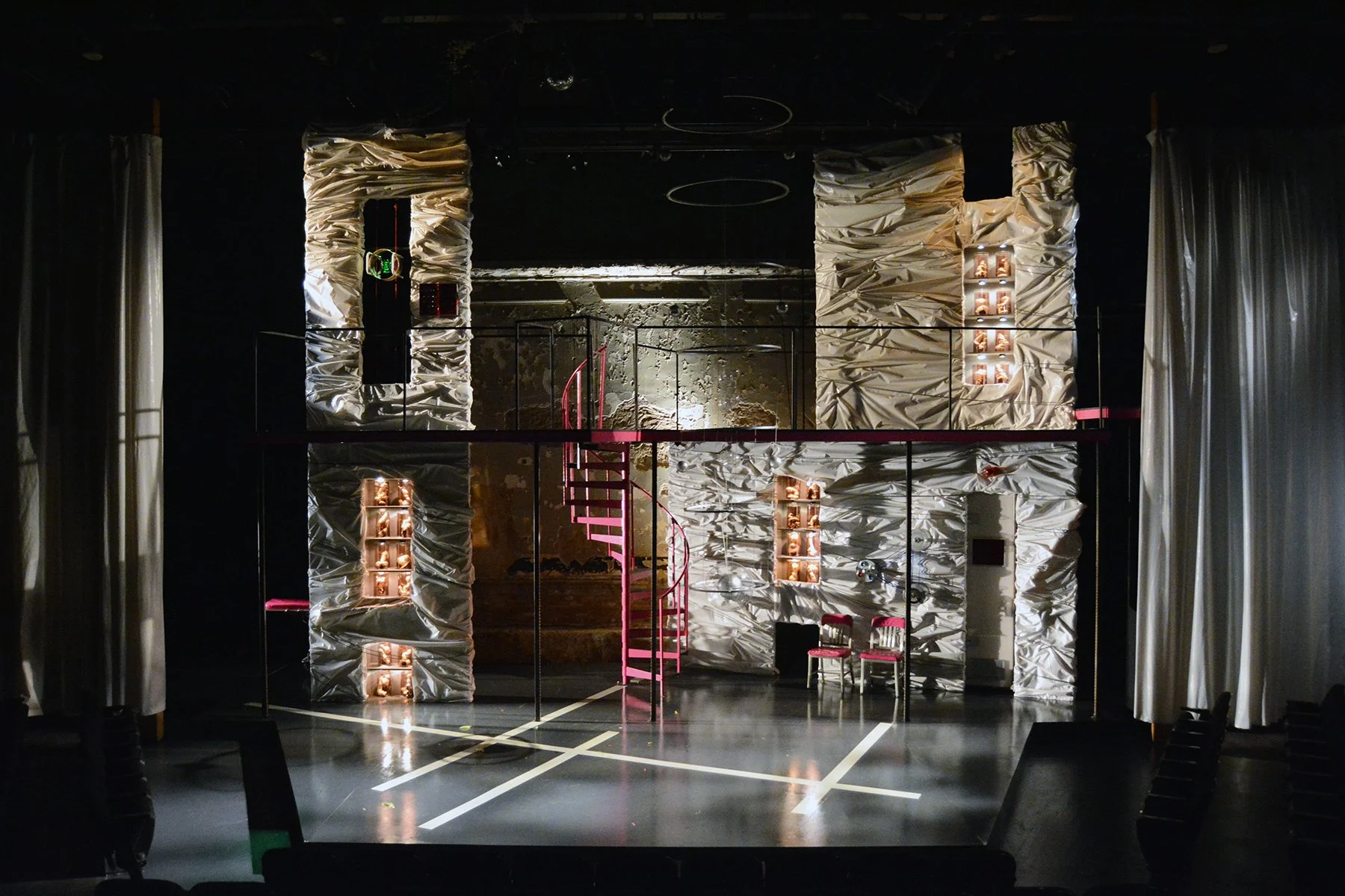 The photo shows a stage with a modern, textured wall backdrop illuminated by warm lighting, a pink spiral staircase at the center, and three chairs on the right side. The stage is framed by white curtains, and there is a black music or sound equipmen