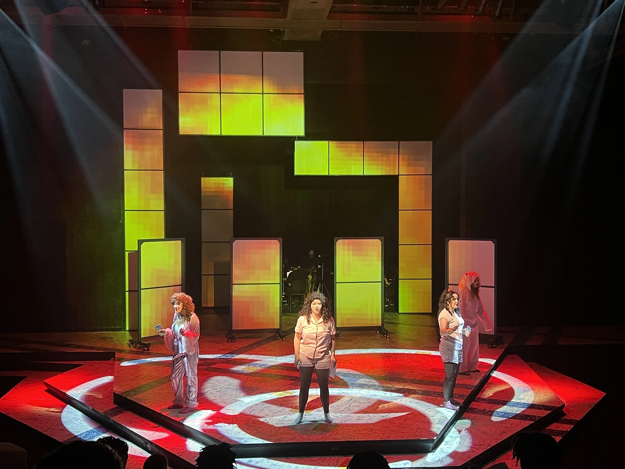 Performers on a stage with vibrant, colorful digital backdrop, some holding props, under dramatic lighting.