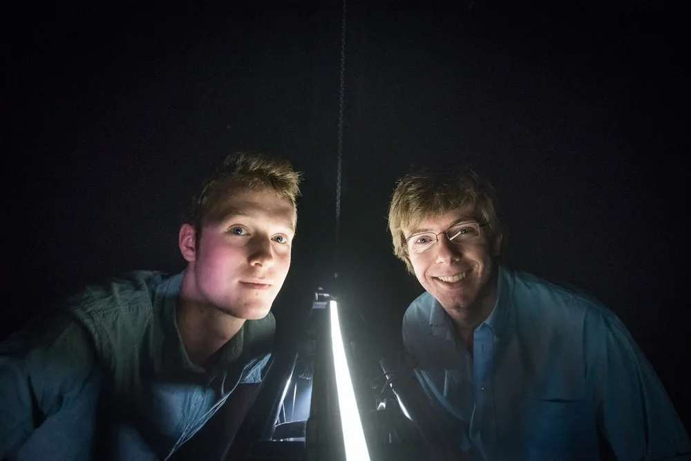Two men smiling and looking into a telescope, with one man wearing glasses, in a dark room with a bright light illuminating the telescope.
