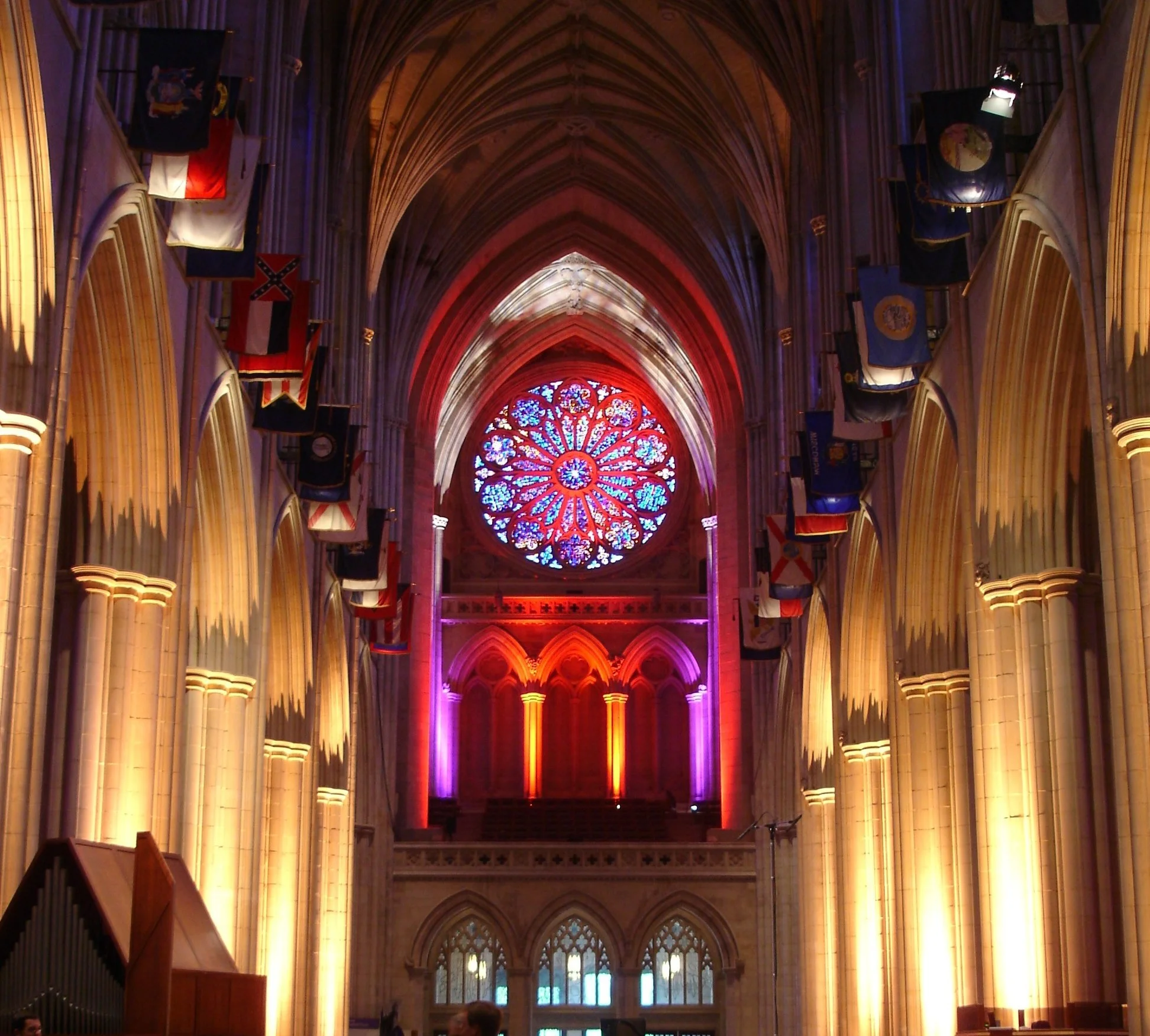 Inside a Gothic cathedral with high vaulted ceilings illuminated in purple and orange lights, stained glass rose window at the end of the nave, and flags hanging on the sides.