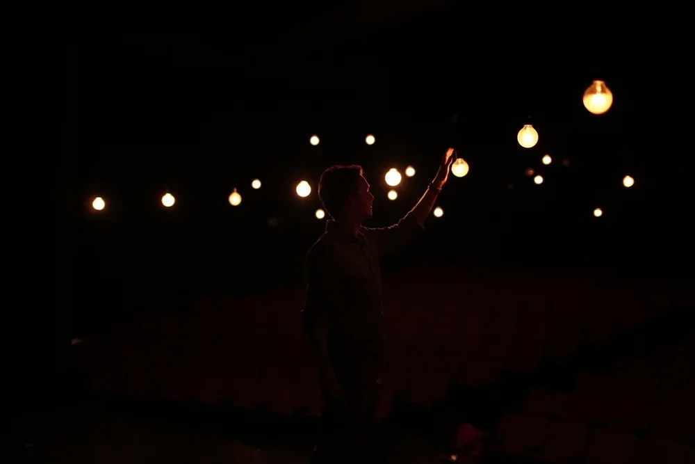 Silhouette of a person touching hanging lamps in a dark room filled with warm lighting.
