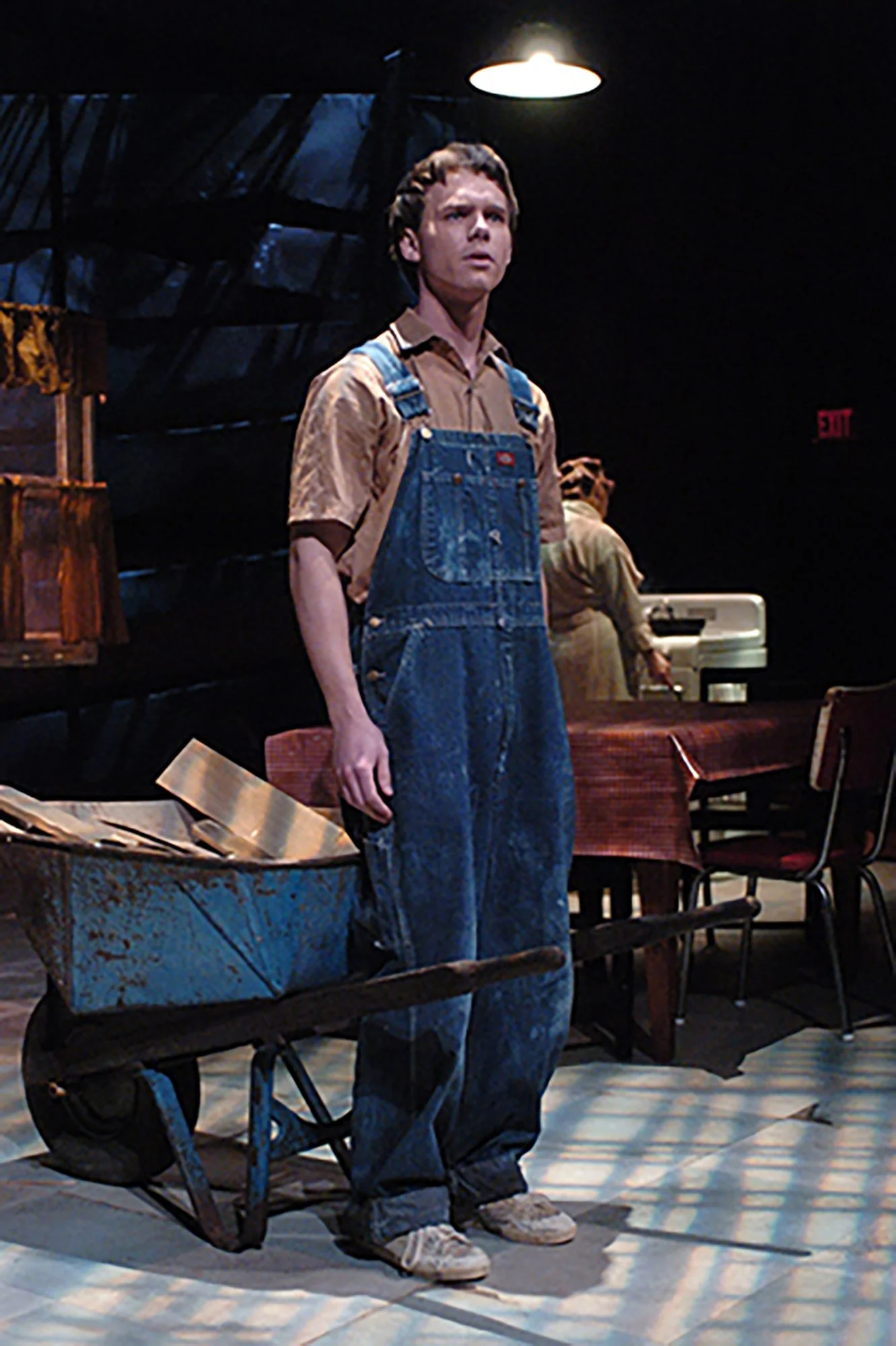 Young man standing on a stage, wearing overalls and a button-up shirt, with a trolley and scattered papers nearby. Dim lighting with a single overhead light illuminating him, as part of a theater or performance setting.