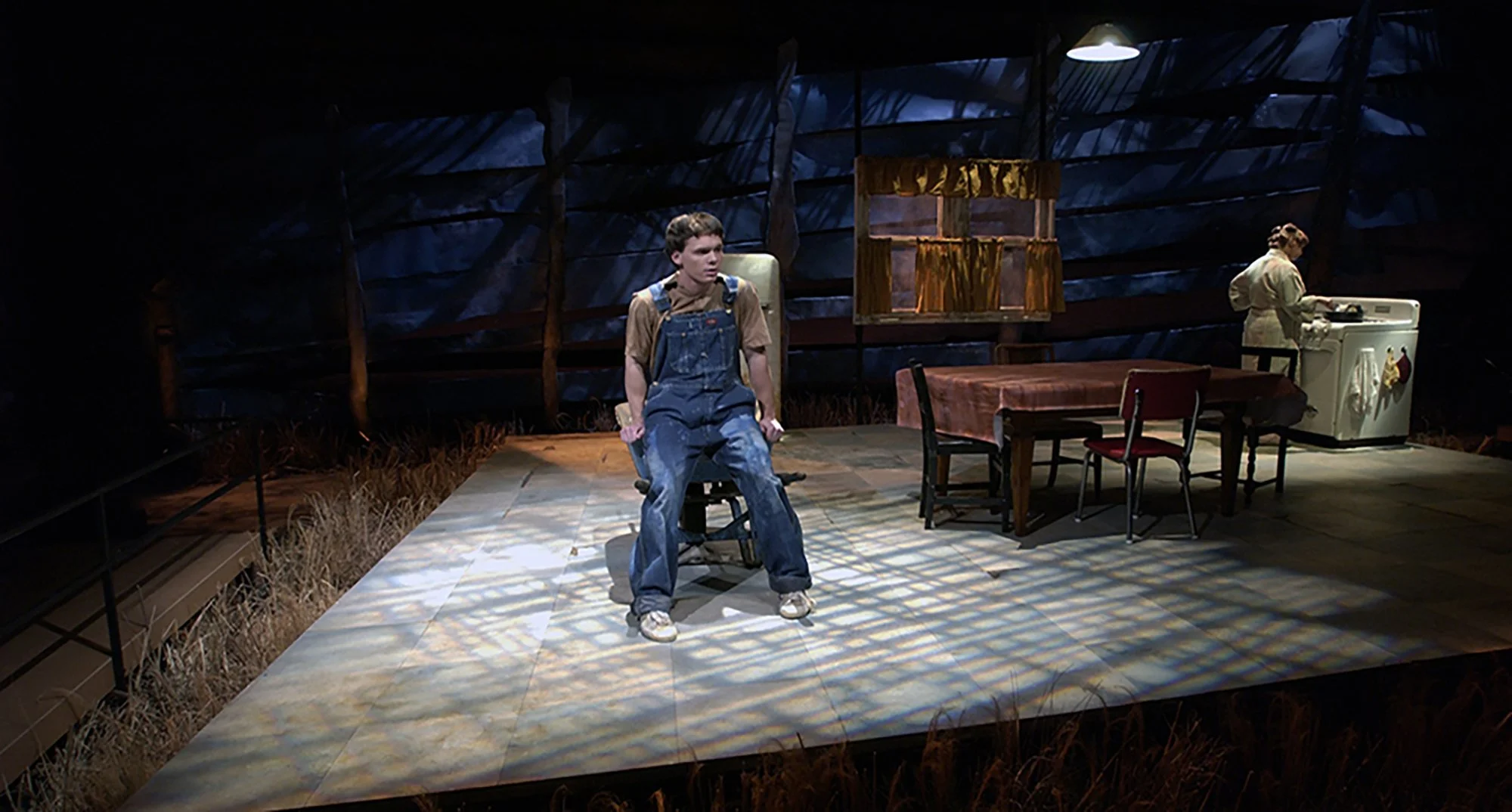Scene from a play or performance showing a young boy sitting in a chair at center stage with a serious expression, wearing overalls and a brown shirt. To the right, a girl is playing a white piano or keyboard. The stage has a rustic setting with a da