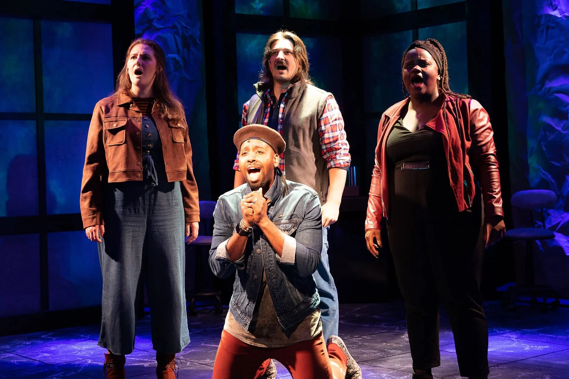 A diverse group of five actors performing on stage with a blue and purple lit background. One man is kneeling in front, one woman on the left, one man in the center, and two women on the right, all expressing strong emotions.