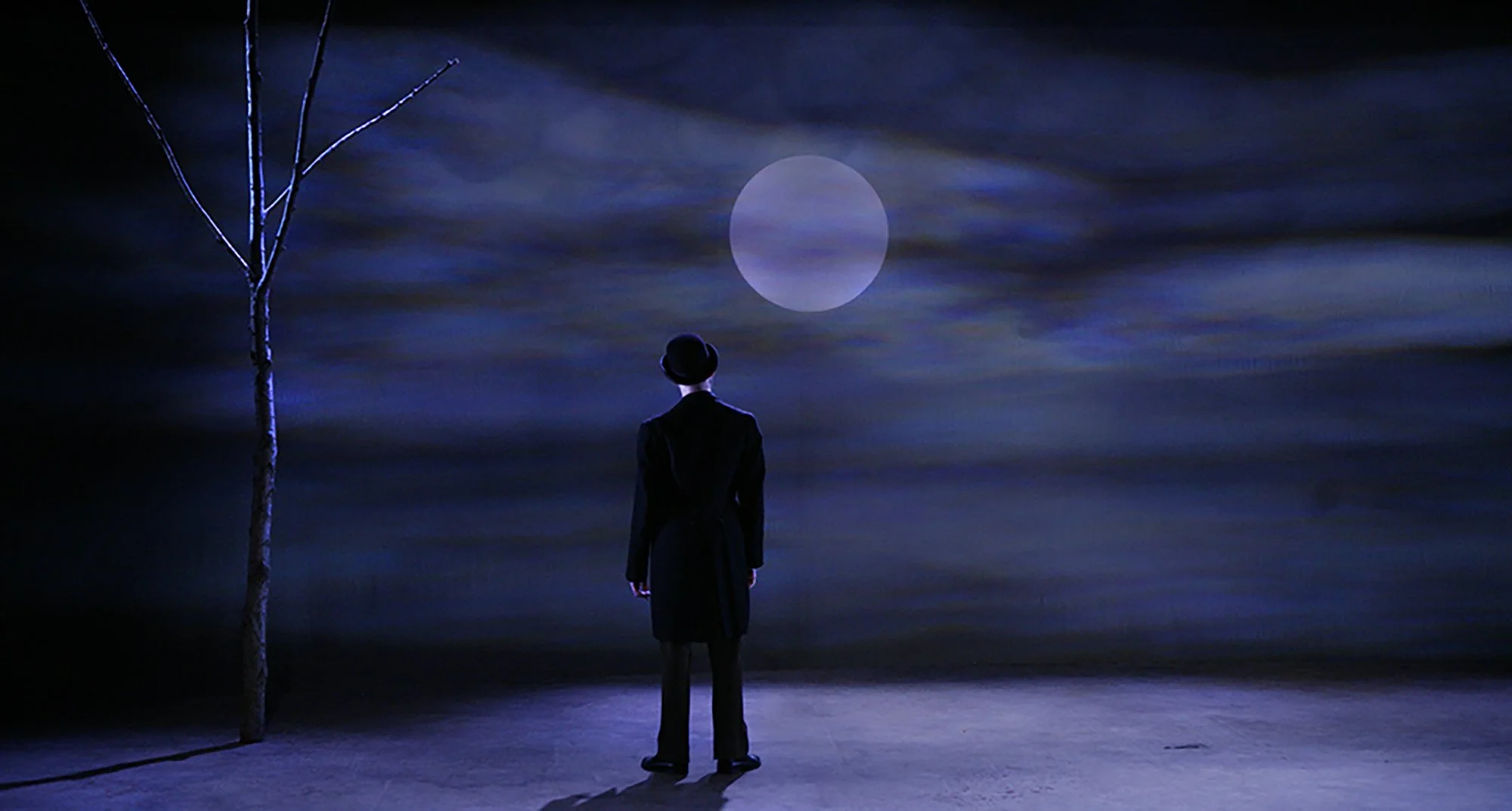 A person in dark clothing and a wide-brimmed hat standing alone on a dark, cloudy landscape, looking at a large, luminous full moon in the night sky, with a barren tree on the left.