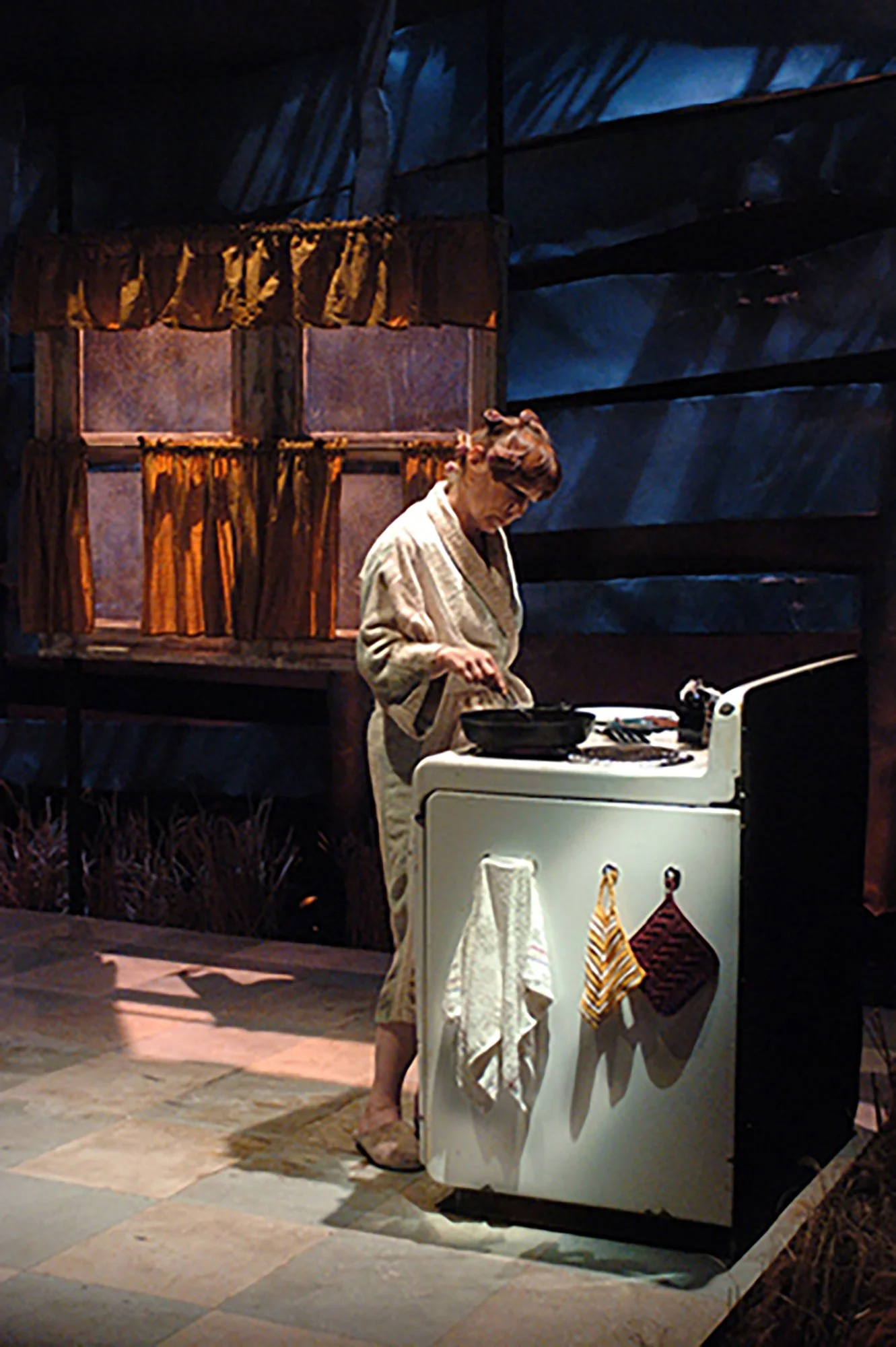 A woman in a bathrobe standing barefoot next to a washing machine with towels hanging on it. Behind her is a dark wall with a window decorated with orange and yellow drapes. She appears to be doing laundry or folding clothes.