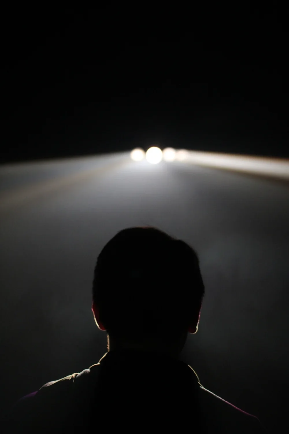 Silhouette of a person's head and shoulders looking at a distant vehicle's bright headlights on a dark road.