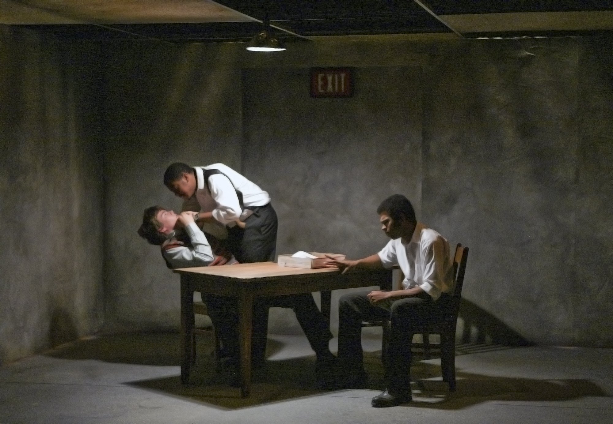 A scene with four people in a dimly lit room with plain walls. Two young men are seated at a table, each holding a pack of cigarettes. Another young man is standing, leaning over the seated man on the left, and appears to be speaking or whispering to