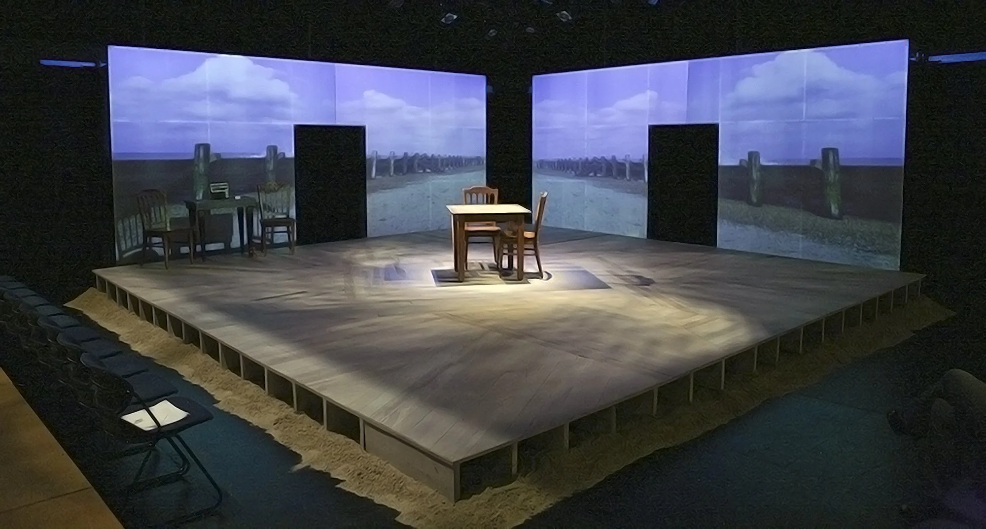 The image shows a theater stage with a set designed to resemble an outdoor scene with a cloudy sky background, wooden chairs, and a table, arranged to depict a seaside or coastal setting.