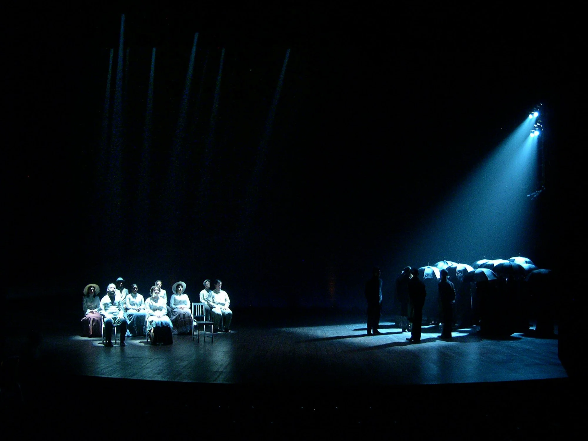 Dimly lit stage with a group of people sitting in chairs on the left and a group standing on the right, with a blue spotlight shining from the right in a theatrical or concert setting.