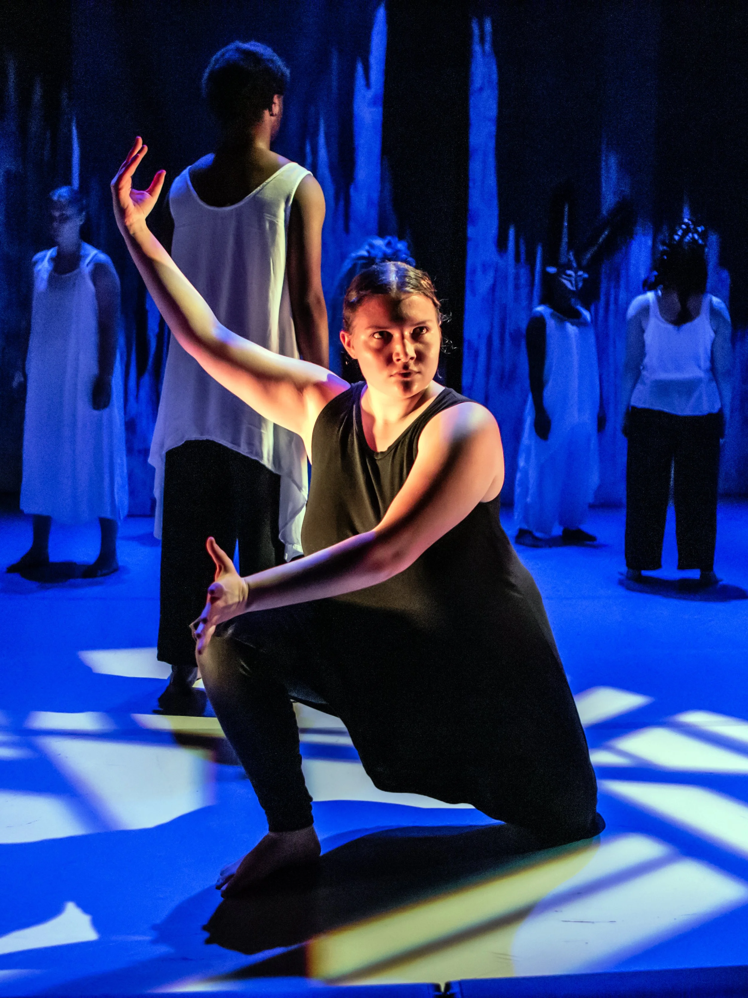 A dancer in a black outfit performs on stage, striking a pose with one arm extended and the other bent at the elbow, surrounded by four others in white shirts and black pants, with a dark background and blue lighting.
