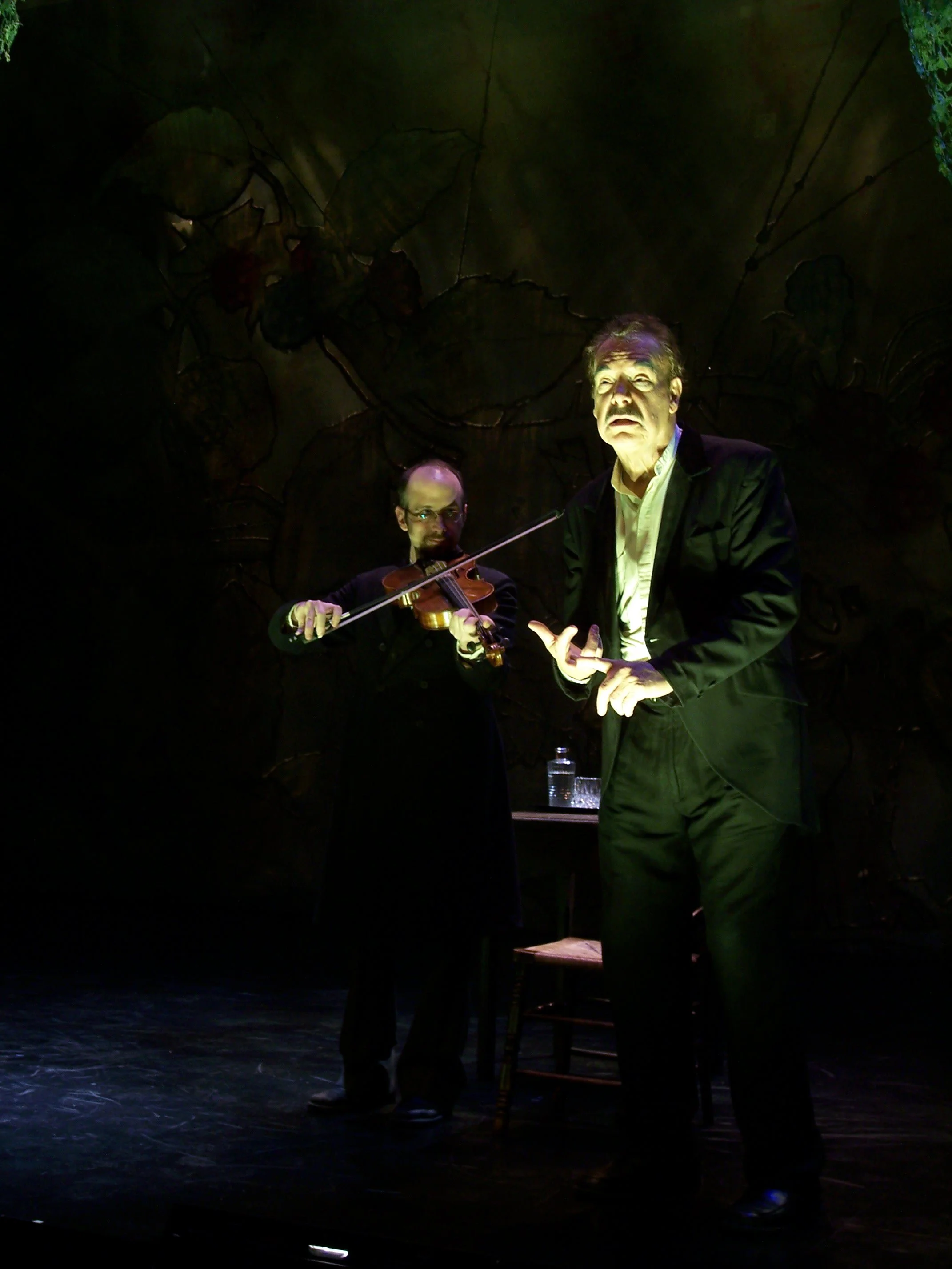Two men on stage, one playing a violin and the other gesturing with his hand, with a dark background featuring a mural of leaves and flowers.