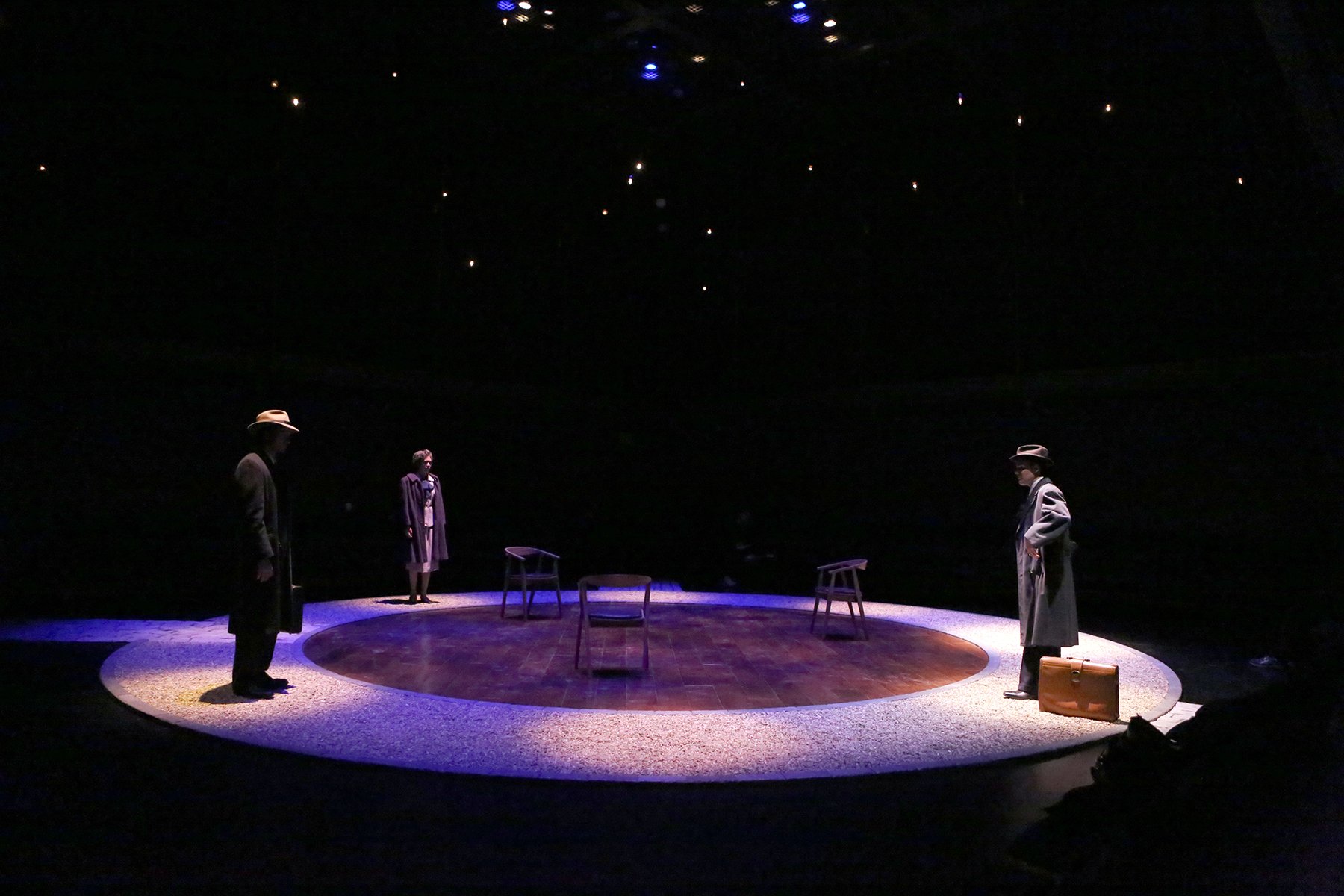 A theatrical scene with three actors standing around a circular stage with chairs and a suitcase. Two of the actors are wearing overcoats and hats, while the third woman is dressed in a purple coat. The stage is dimly lit with a dark background and s