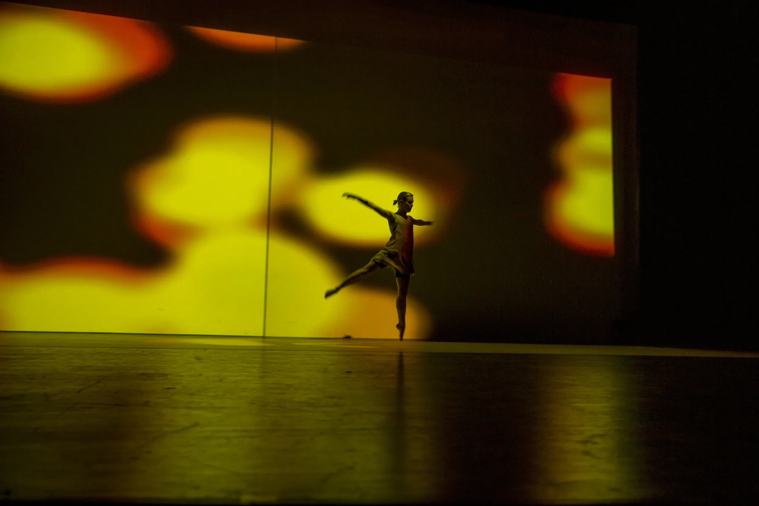 A dancer performing on stage with a colorful abstract background projected behind her, illuminated in yellow light.