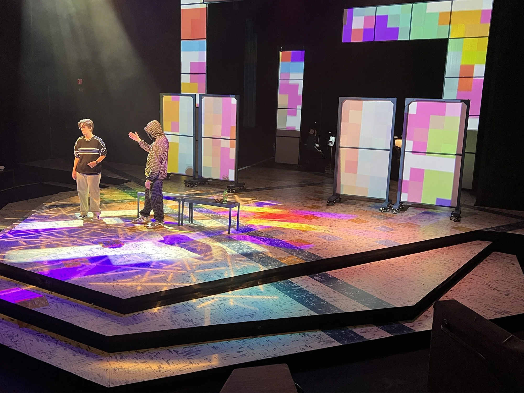 Two people on a stage with colorful digital screens and lighting effects, one appears to be speaking or gesturing while the other stands with hands in pockets.