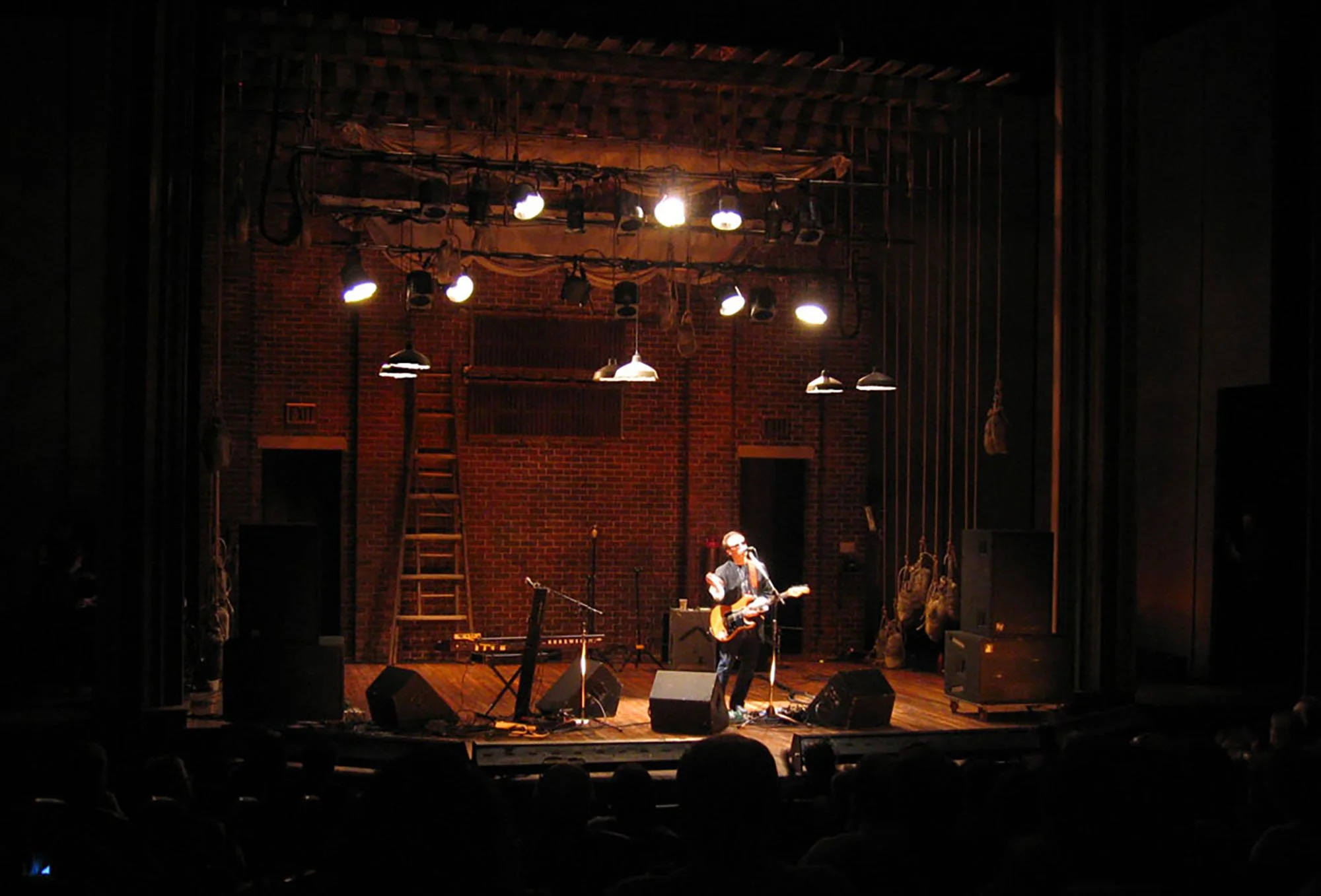 A musician performs on stage in a dimly lit venue with brick walls. The performer is playing an acoustic guitar and singing into a microphone, with stage lights overhead.