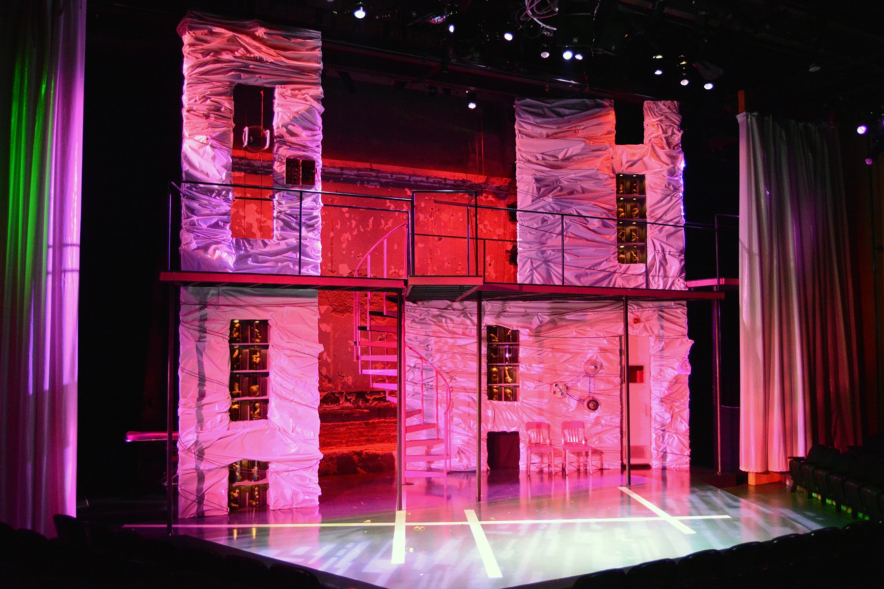 Stage decorated with crinkled white fabric, illuminated by pink and purple lights. Multiple shelves and curtains are visible, with a spiral staircase in the center. The scene appears to be set for a theatrical performance or a play.