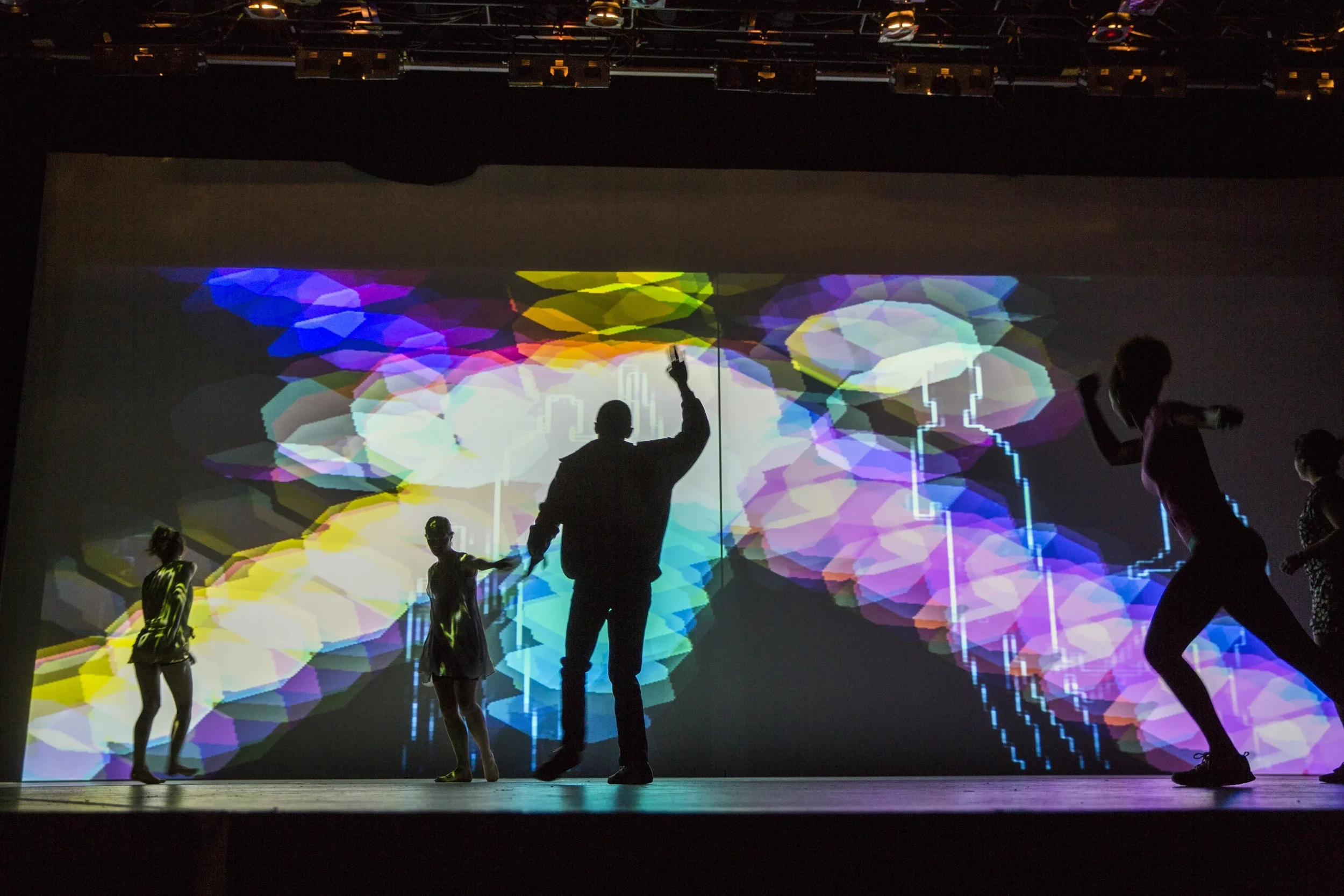 Silhouettes of people on stage with vibrant, colorful, geometric light projections in the background.