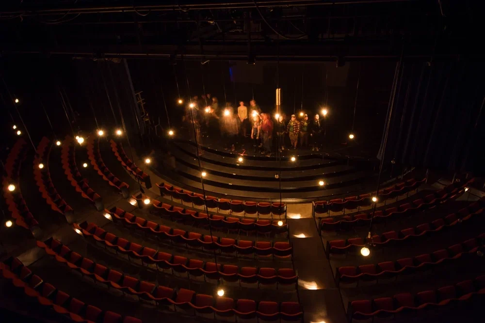 Empty theater with red seats, lit by hanging lights, with a small group of people on stage.